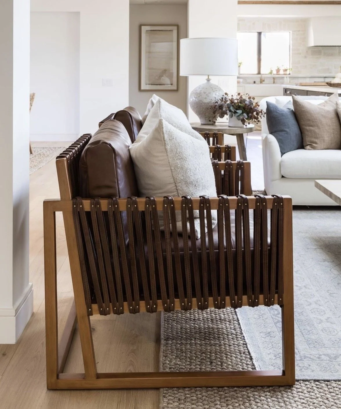Living room with brown leather and wooden framed couch, white and beige pillows, side table with lamp and vase, and white sofa in background.