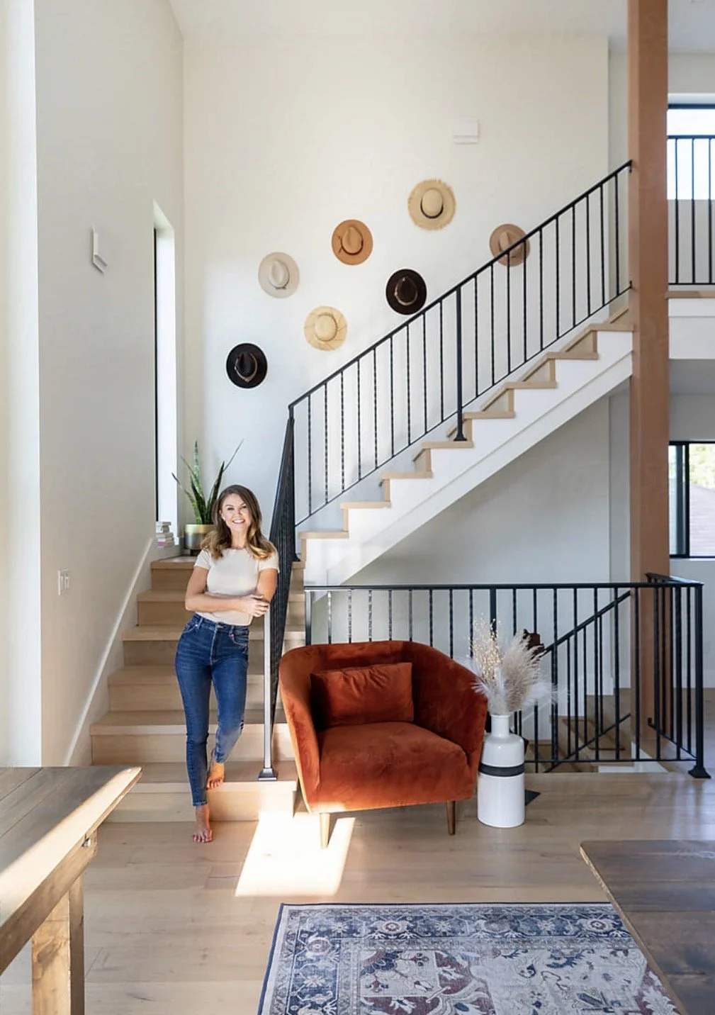 A woman in casual clothing standing in a modern living room with a staircase decorated with hats on the wall, a rust-colored armchair, a white vase with dried flowers, and a rug.