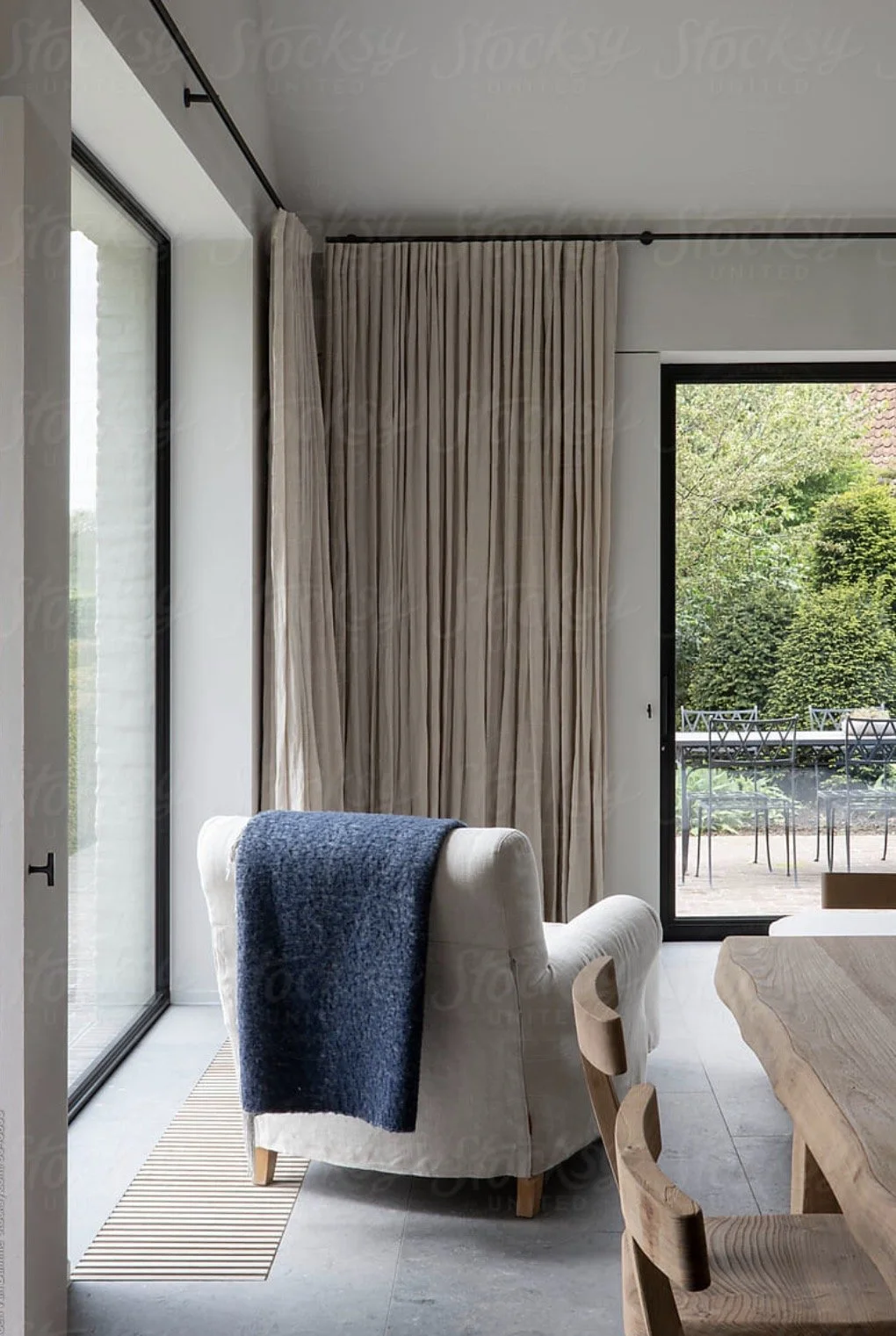 A cozy living room with a white armchair having a blue blanket draped over it, next to large sliding glass doors with beige curtains, leading to an outdoor patio with a table and chairs, and greenery visible outside.