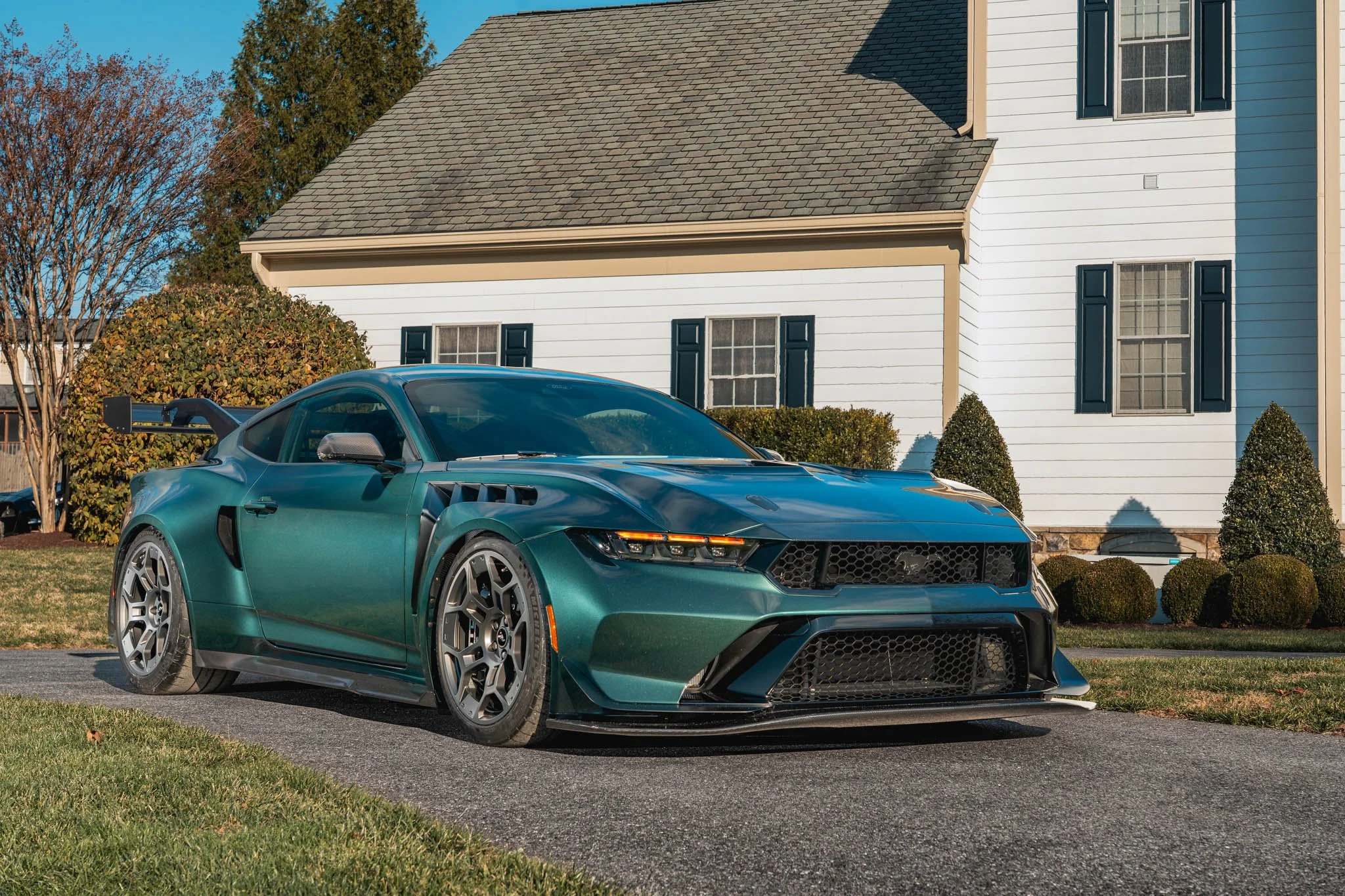 A green sports car parked in front of a white house with blue shutters and a gabled roof, surrounded by bushes and trees on a clear day.