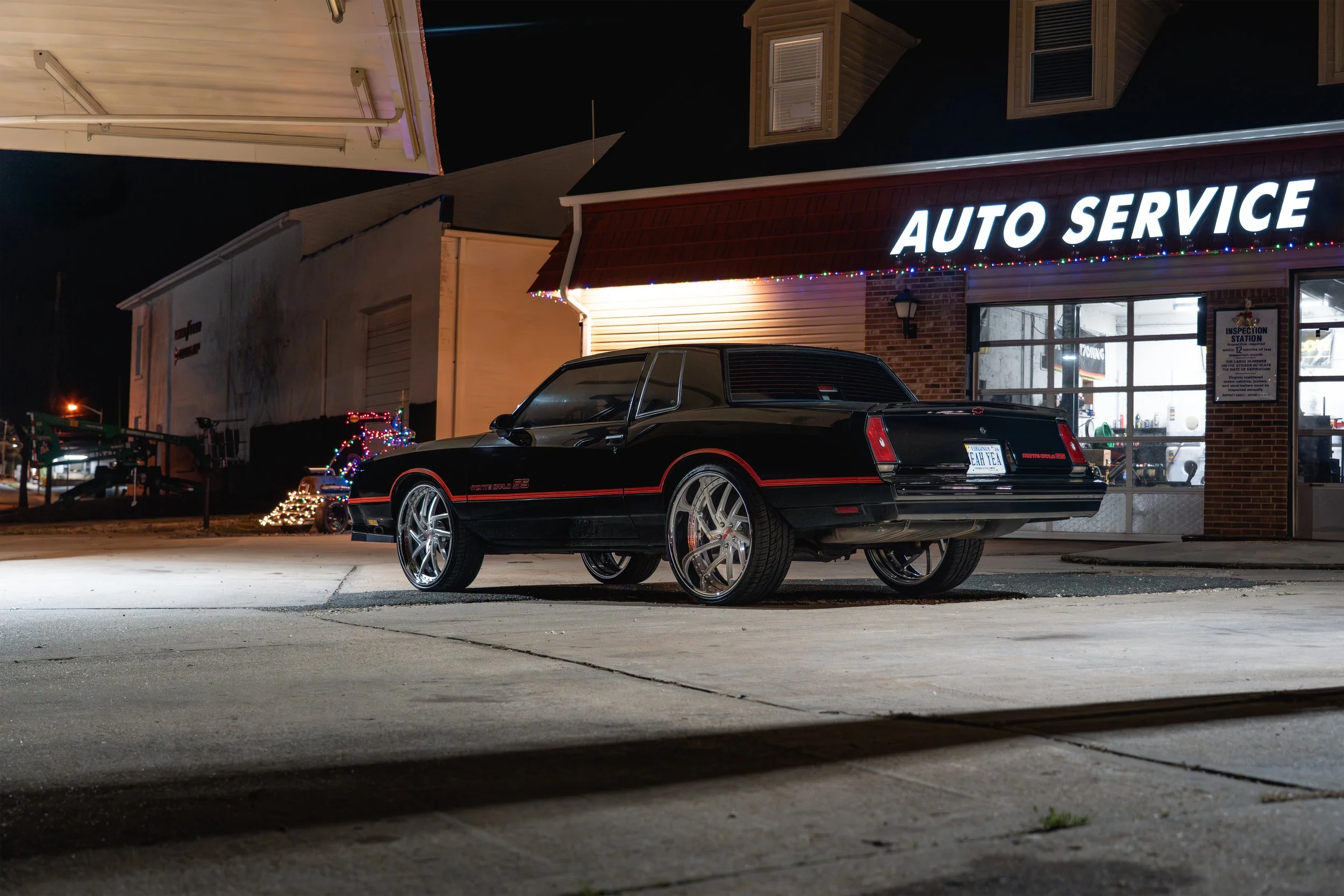 A black vintage car with large chrome wheels parked outside an auto service shop at night. The shop has a bright illuminated sign and holiday lights along the roofline. The surrounding area is dark with a few buildings and lights in the distance.