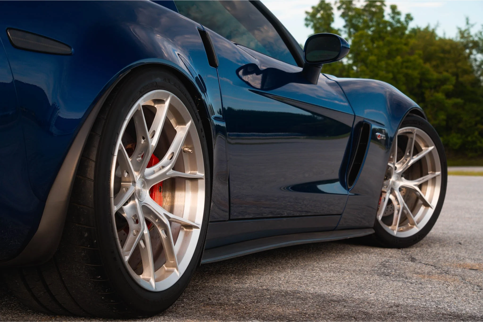 Close-up view of a blue sports car showing its front left wheel, side mirror, and part of the hood, with green trees in the background.