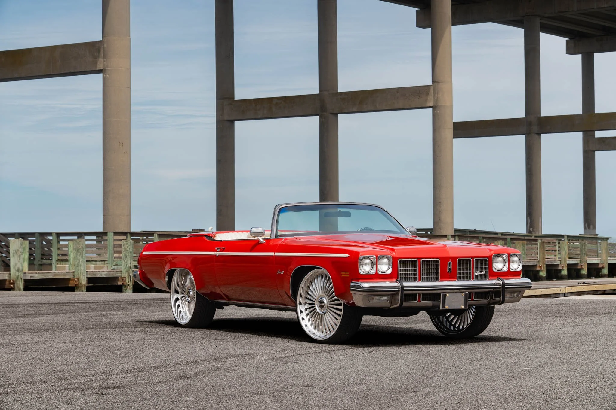 A vintage red convertible car parked on a paved surface near a wooden fence. In the background, there is a large concrete bridge with tall support columns and a clear sky.