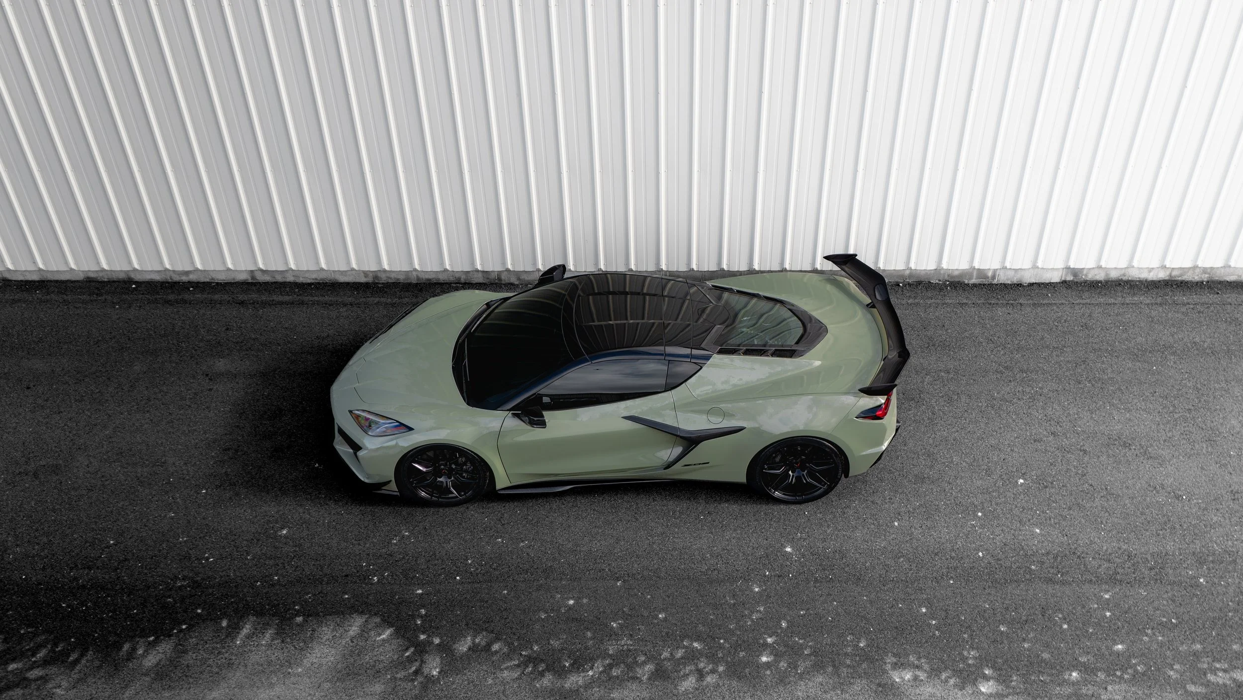 Top-down view of a light green sports car parked on a dark asphalt surface beside a corrugated metal wall.