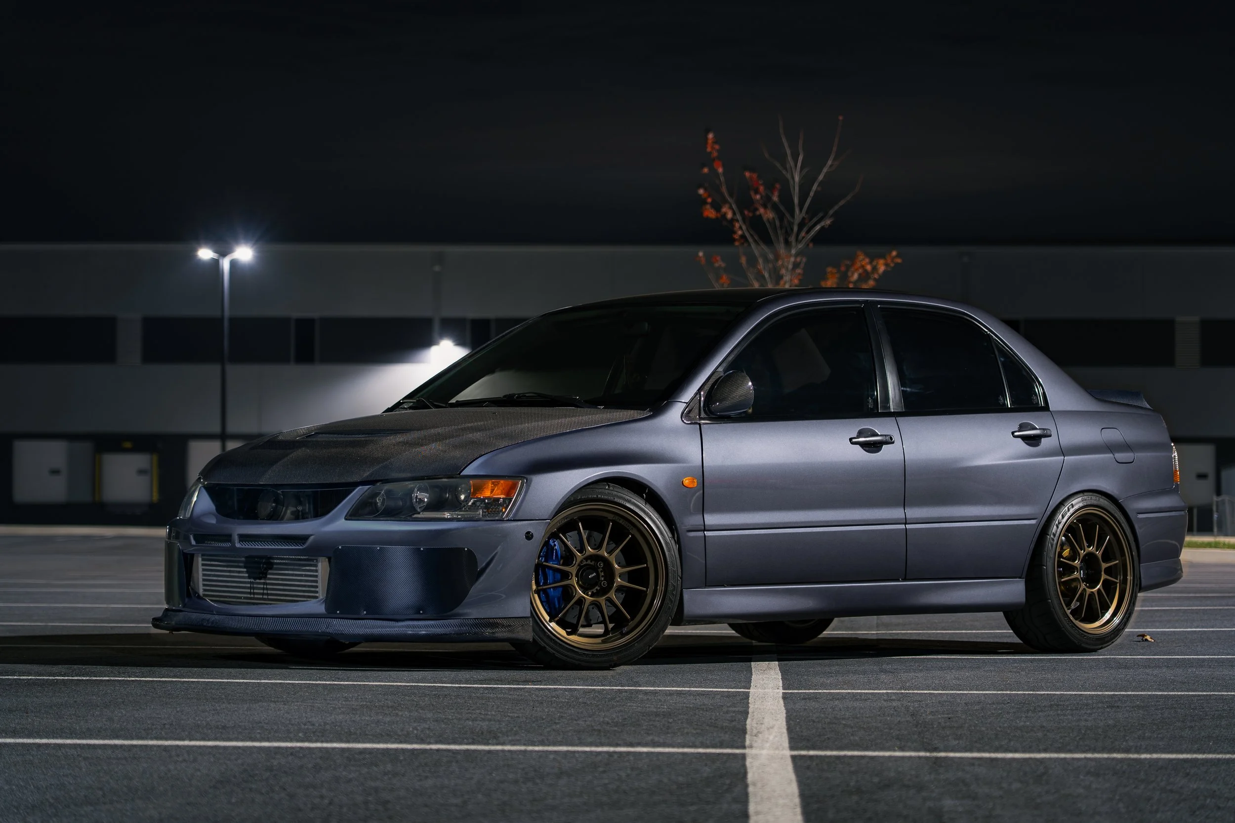 A silver modified sports car parked in a well-lit parking lot at night, with a dark building and a small tree with red leaves in the background.