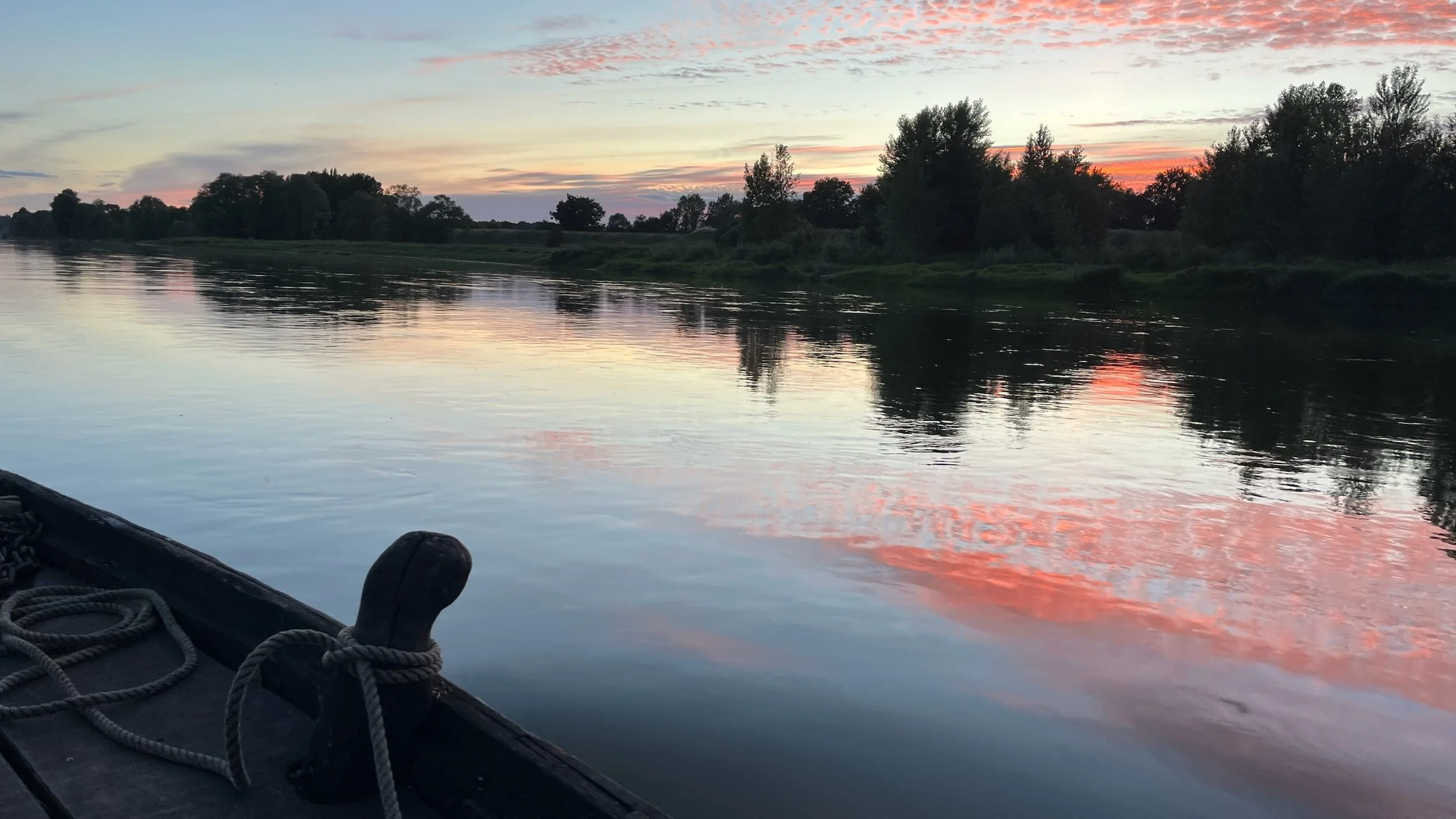 Coucher de soleil sur une rivière calme avec des arbres en silhouette à la rive, vu depuis un bateau avec des cordages en premier plan