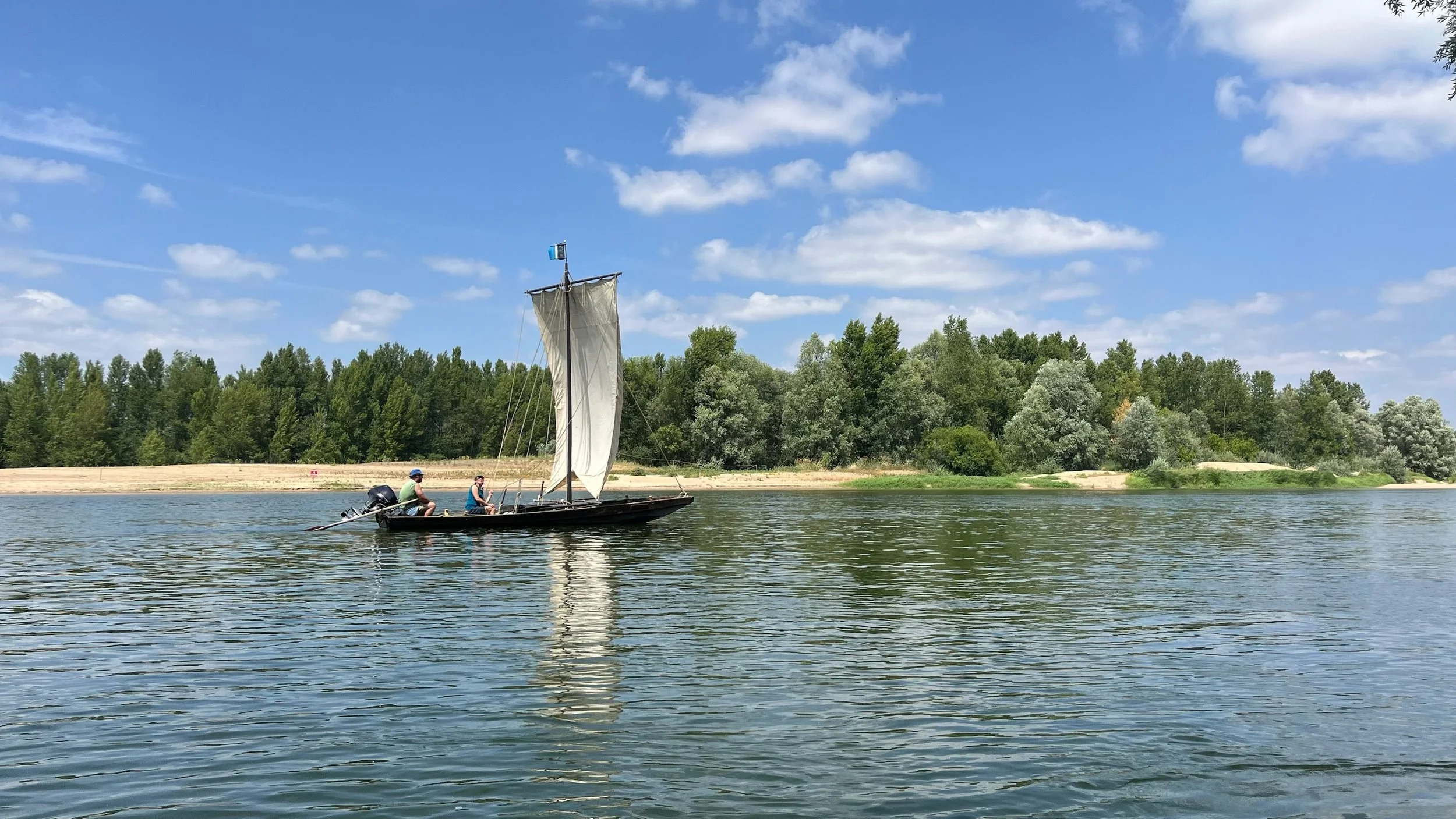 Un petit voilier avec deux personnes à bord navigue sur une rivière, avec une forêt dense et un ciel bleu avec quelques nuages en arrière-plan.