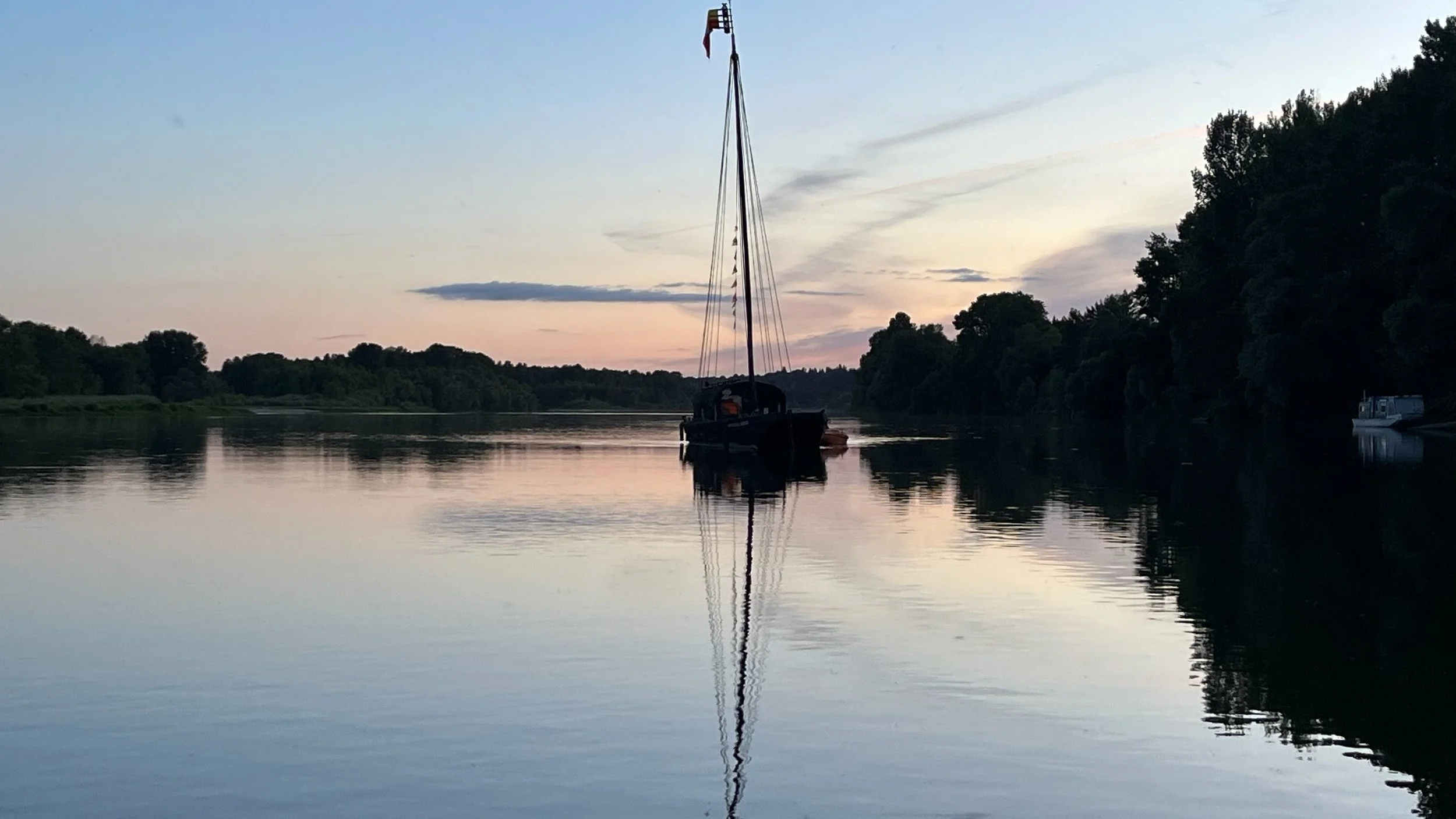 Un bateau à voile sombre flottant sur une rivière calme au coucher du soleil, entouré d'arbres en silhouette et un ciel aux couleurs pastel.