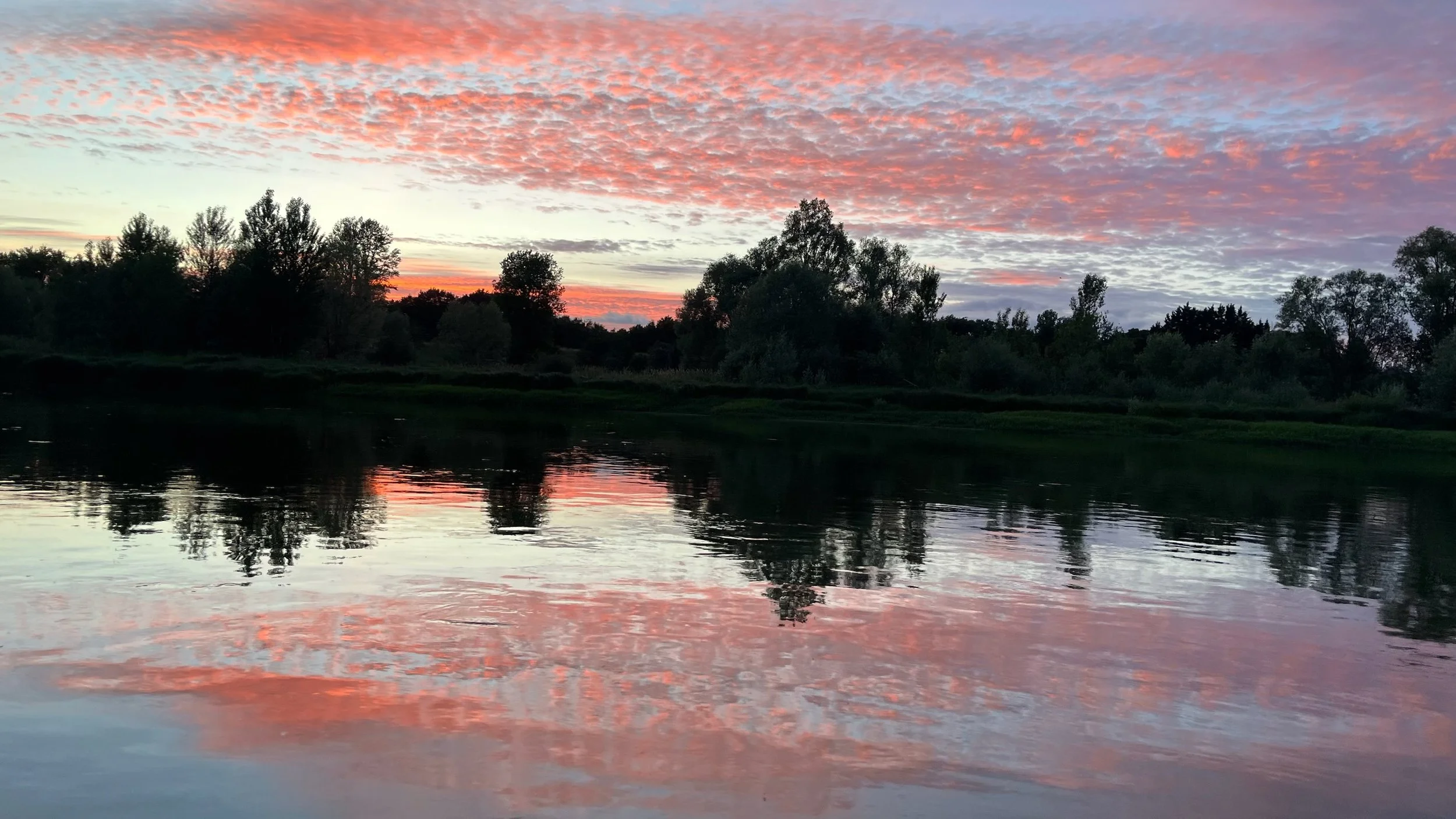 Coucher de soleil sur une rivière avec des arbres en silhouette et un ciel avec des nuages roses et orange.