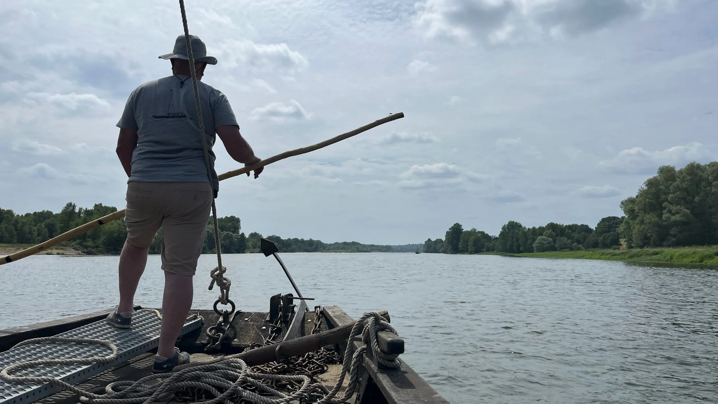Un homme debout sur un bateau en rivière, tenant une longue perche, portant un chapeau, un t-shirt gris et un short beige, avec des arbres en arrière-plan sous un ciel nuageux.