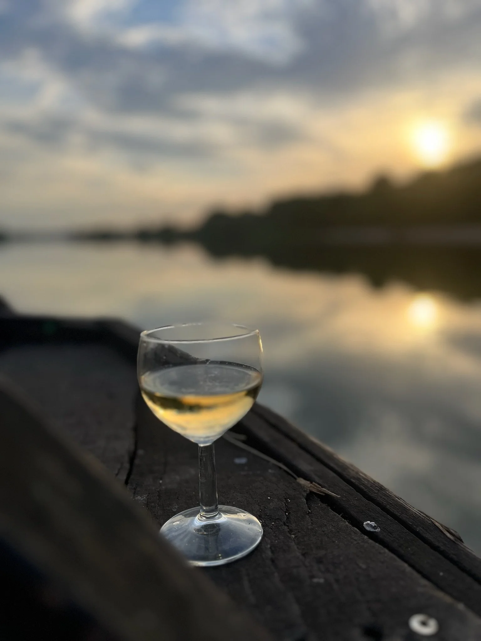 Verre de vin blanc placé sur une surface en bois près d'un lac au coucher du soleil, avec un ciel nuageux.