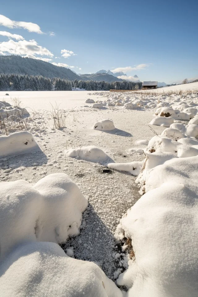Winter am Ufer des Geroldsee, Schnee und Eis dominieren das Bild während blauer Himmel im Hintergrund die Zugspitze hervortreten lässt.