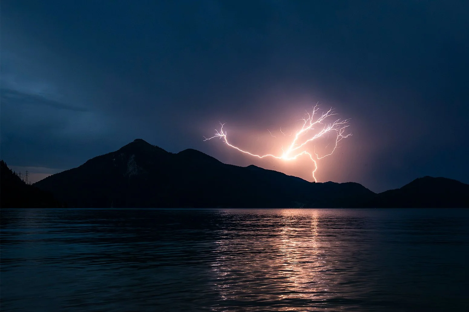 Ein Gewitterblitz am dunklen Himmel neben dem Jochberg in den bayrischen Voralpen, das Licht spiegelt sich im Wasser des Walchensee.