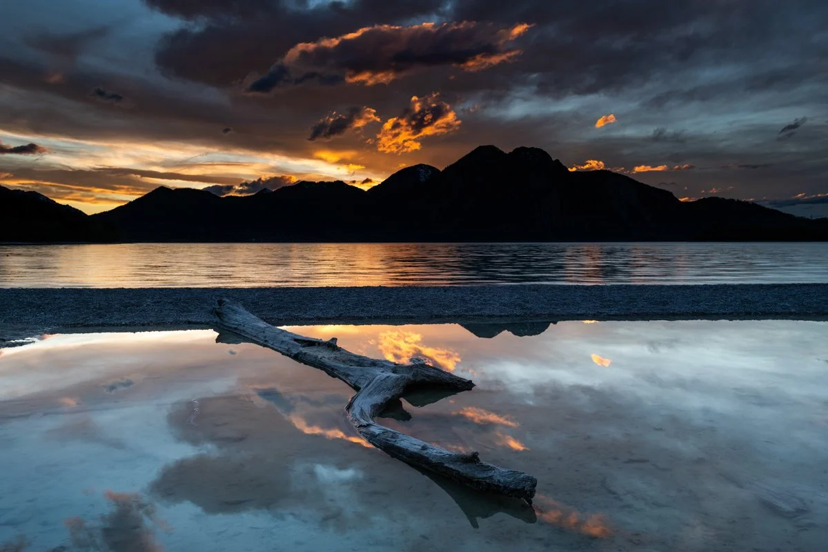 Abendrot am Ufer des Walchensee. Im Vordergrund ein alter Baumstamm am Ufer des See und im Hintergrund der Herzogstand mit einem spektakulären Sonnenuntergang.