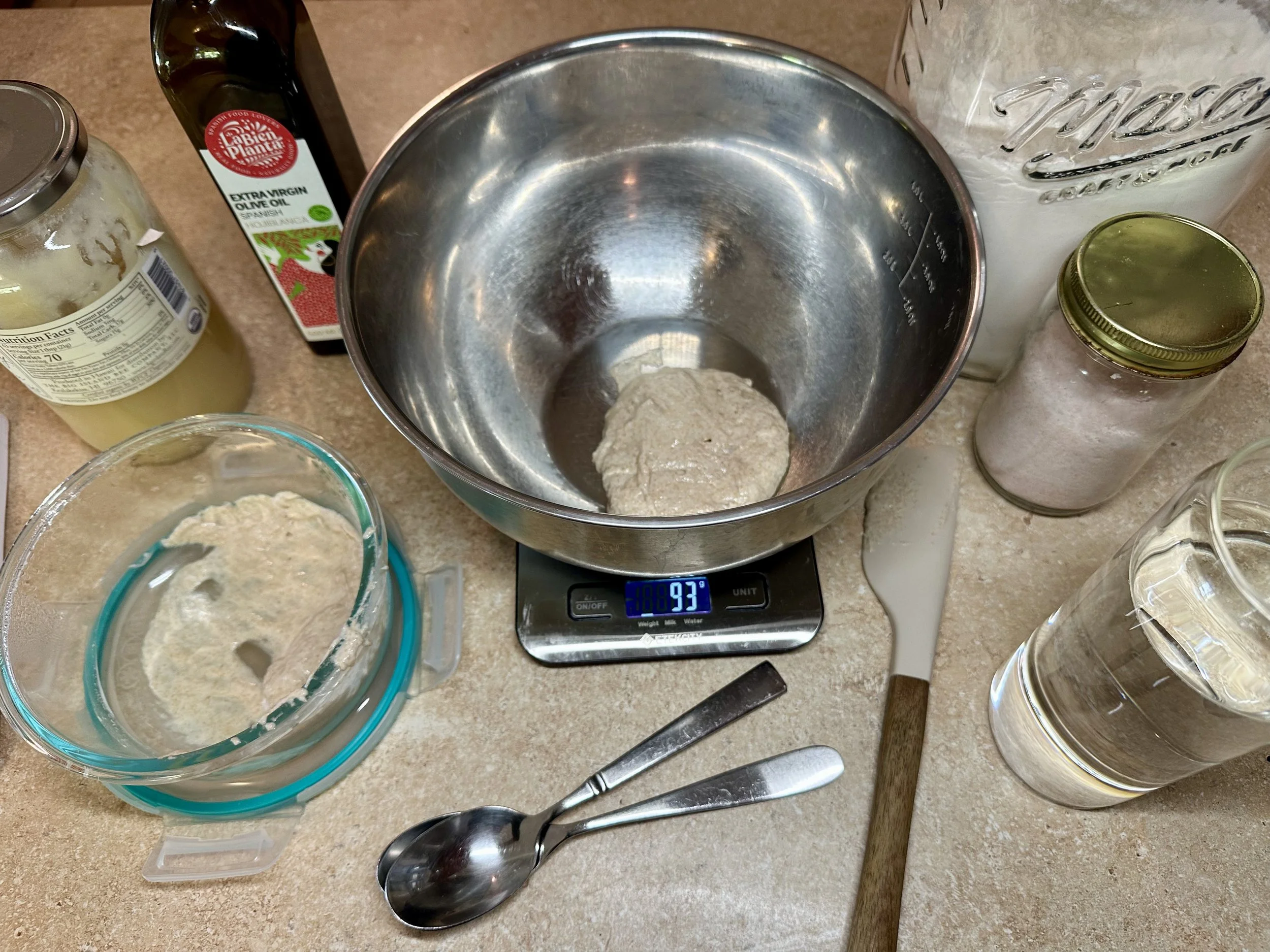 Sourdough sandwich bread ingredients on a kitchen counter with starter, flour, olive oil, honey, water, and mixing bowl on a scale