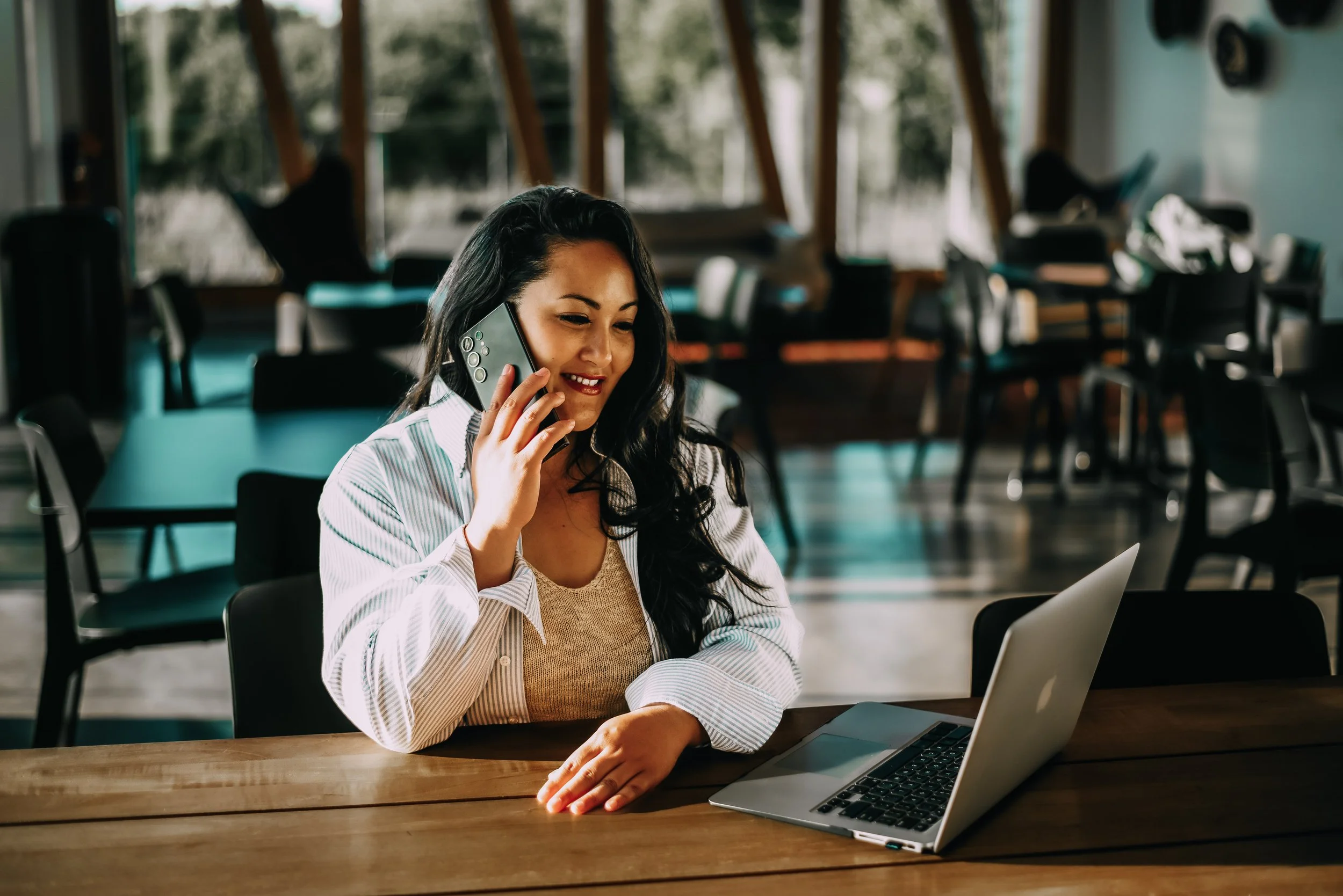 A woman with long black hair, wearing a white striped shirt and brown top, smiling while talking on a mobile phone at a wooden table with a silver laptop in front of her, in a bright, modern indoor space with large windows and black chairs.