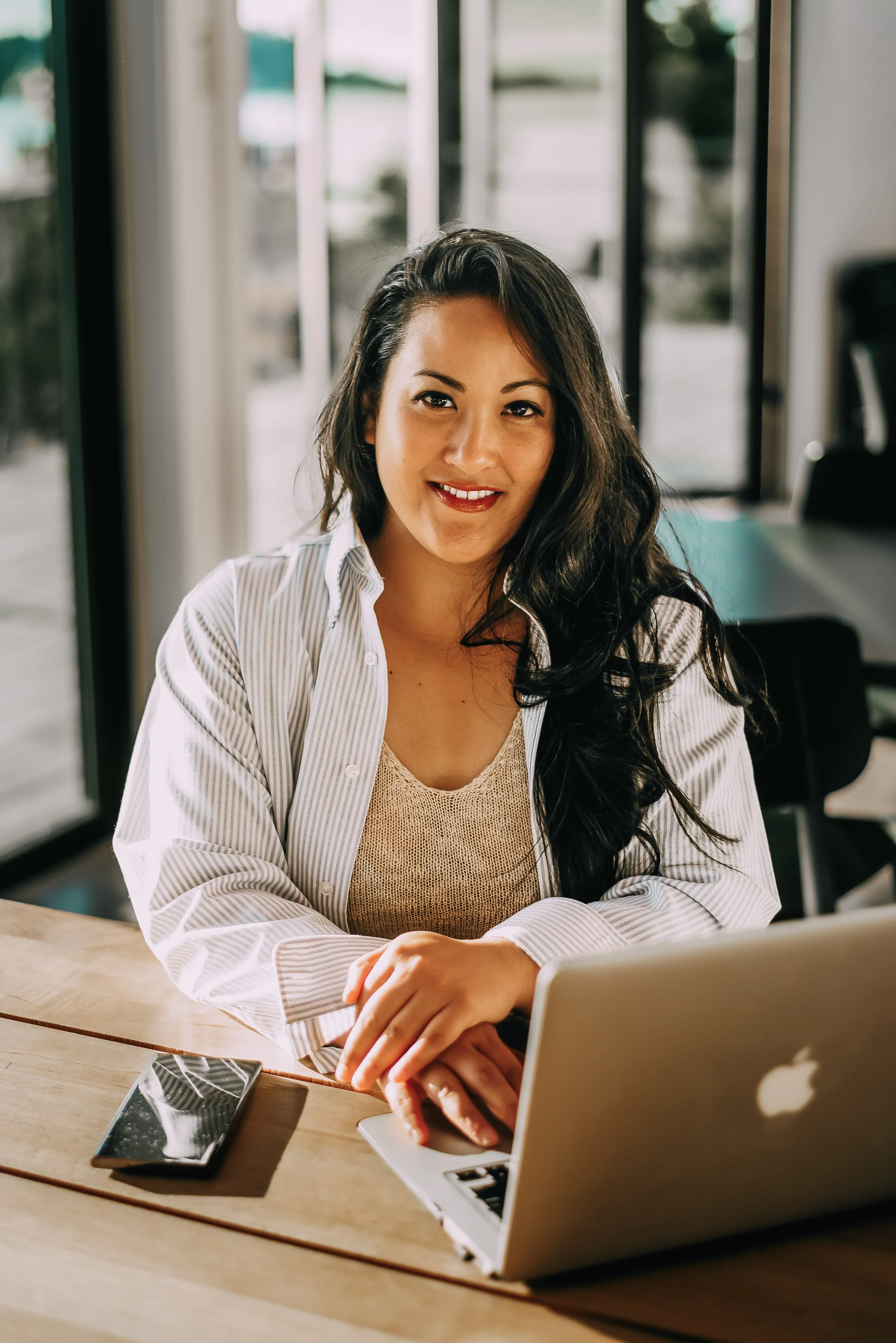 A woman with long dark hair and a bright smile sitting at a wooden table in a modern office or café, using a silver laptop, with a smartphone placed on the table and large windows in the background.