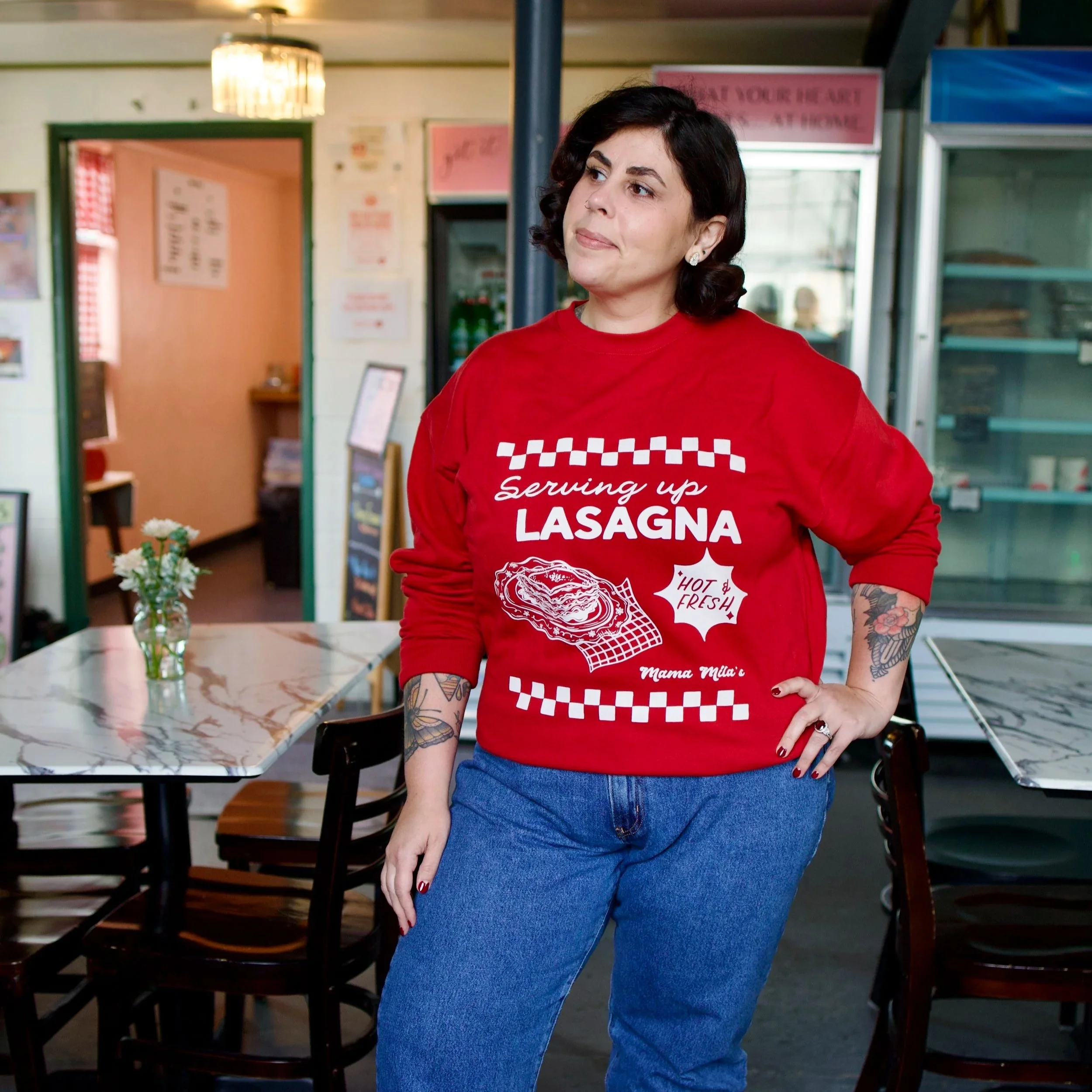 Mila in a red Mama Mila’s sweatshirt featuring a vintage Italian diner-inspired white graphic with “Serving Up Lasagna” lettering, an illustrated plate of lasagna, “Hot & Fresh!” text, and retro check
