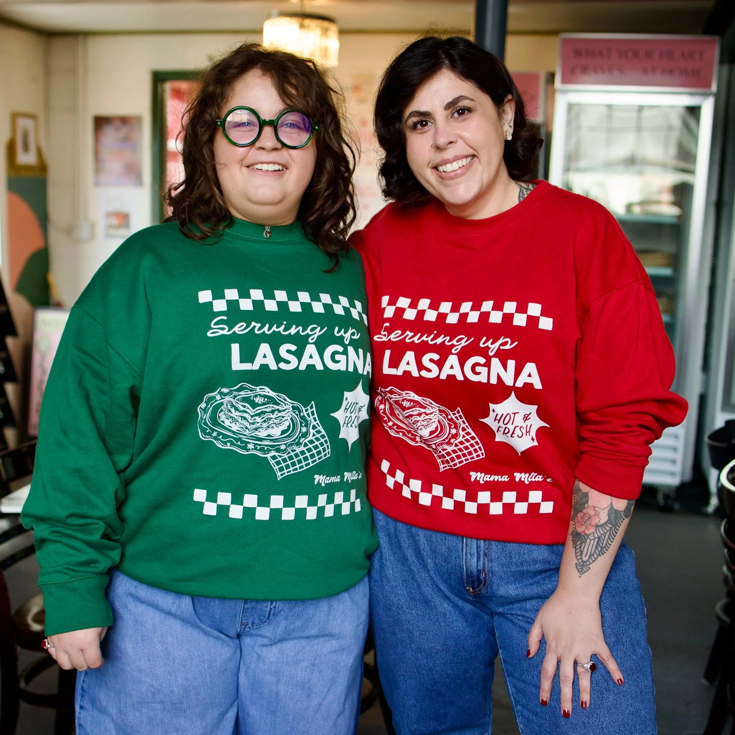Two women modeling Mama Mila’s Serving Up Lasagna crewneck sweatshirts in festive red and green, featuring retro Italian diner graphics, white checkerboard accents, and lasagna illustrations.