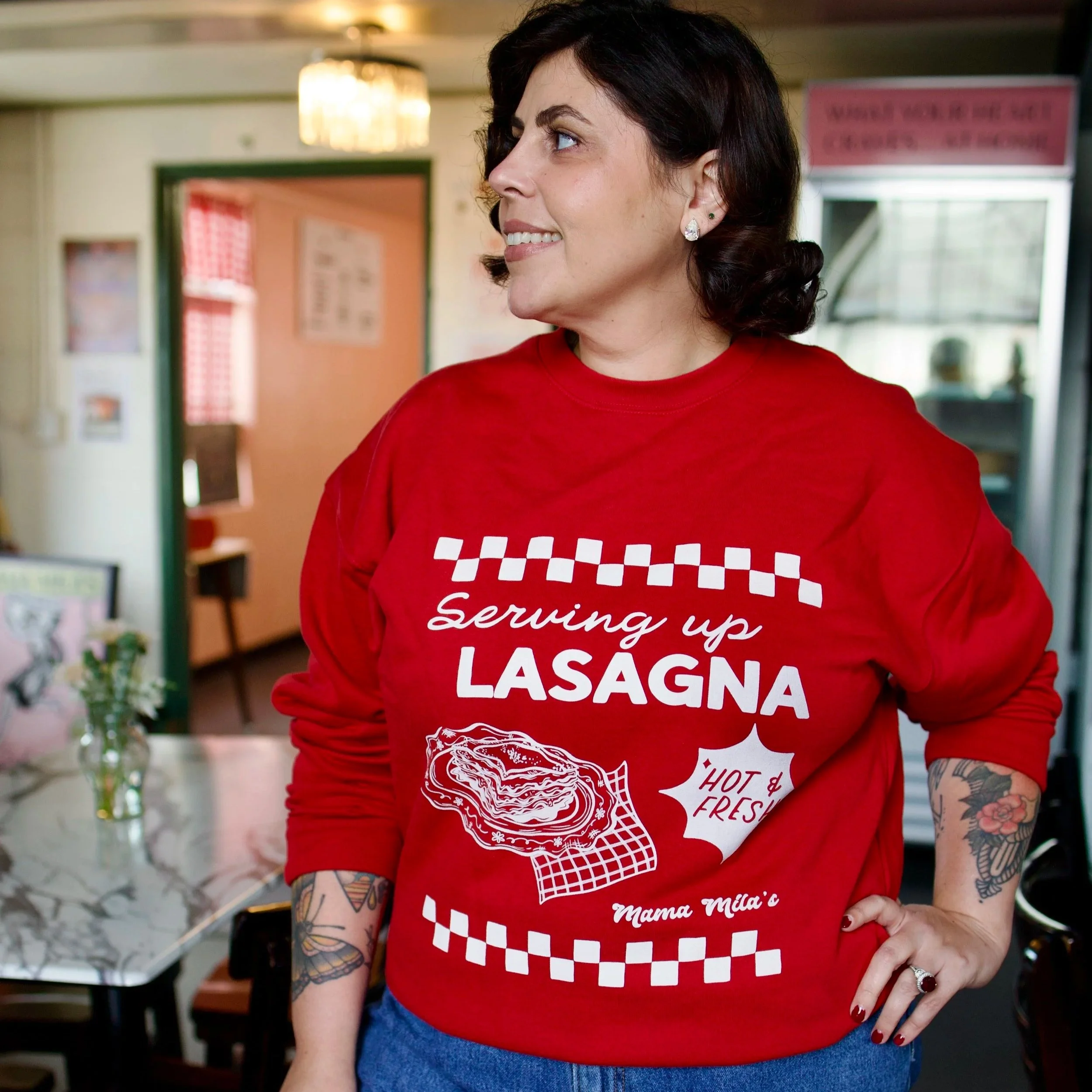 Mila in a red Mama Mila’s sweatshirt featuring a vintage Italian diner-inspired white graphic with “Serving Up Lasagna” lettering, an illustrated plate of lasagna, “Hot & Fresh!” text, and retro check