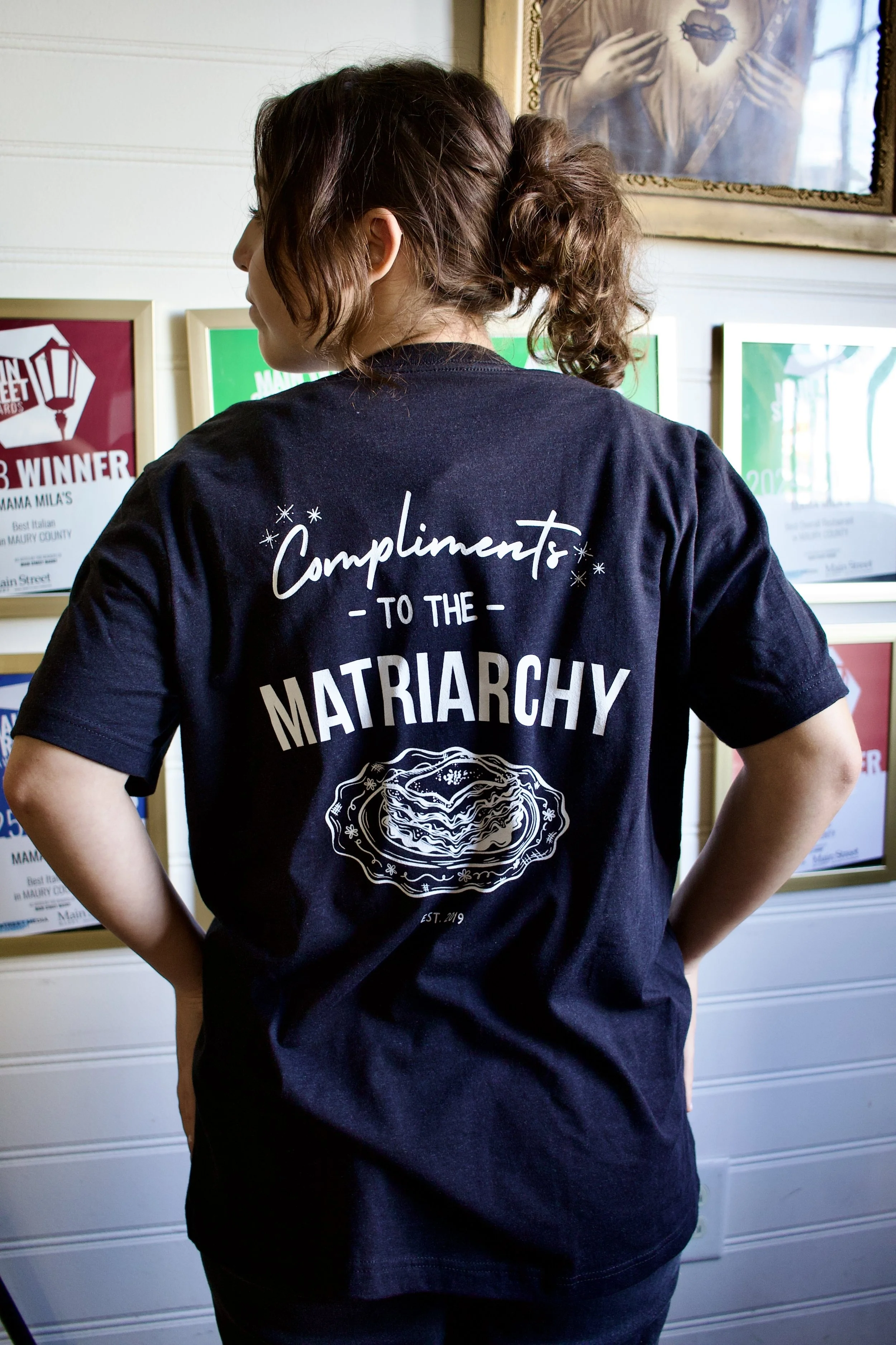 Compliments to the Matriarchy with a piece of lasagna on a vintage plate on the back of a black t-shirt in front of a white wall of awards.