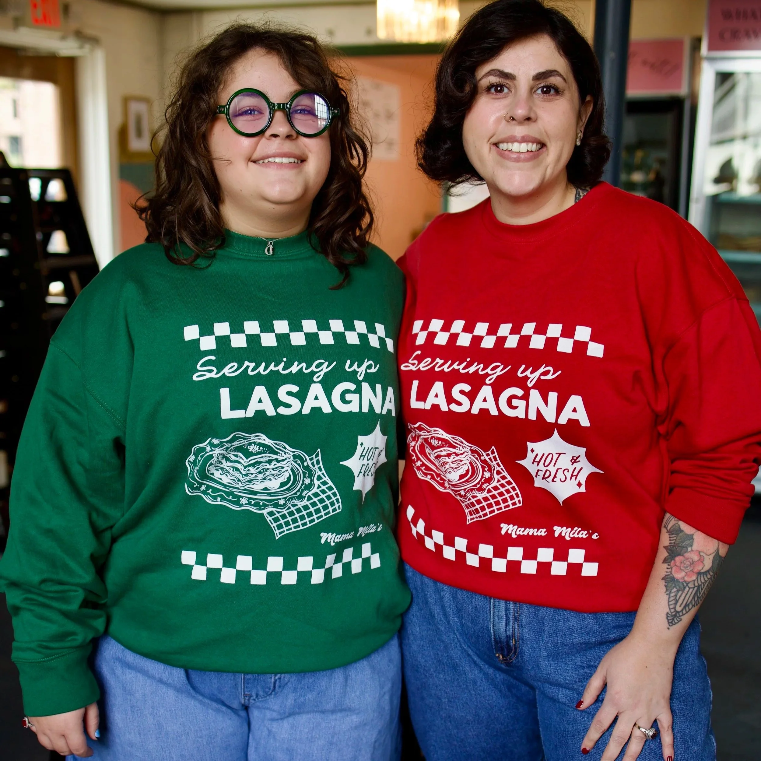 Two women modeling Mama Mila’s Serving Up Lasagna crewneck sweatshirts in festive red and green, featuring retro Italian diner graphics, white checkerboard accents, and lasagna illustrations.