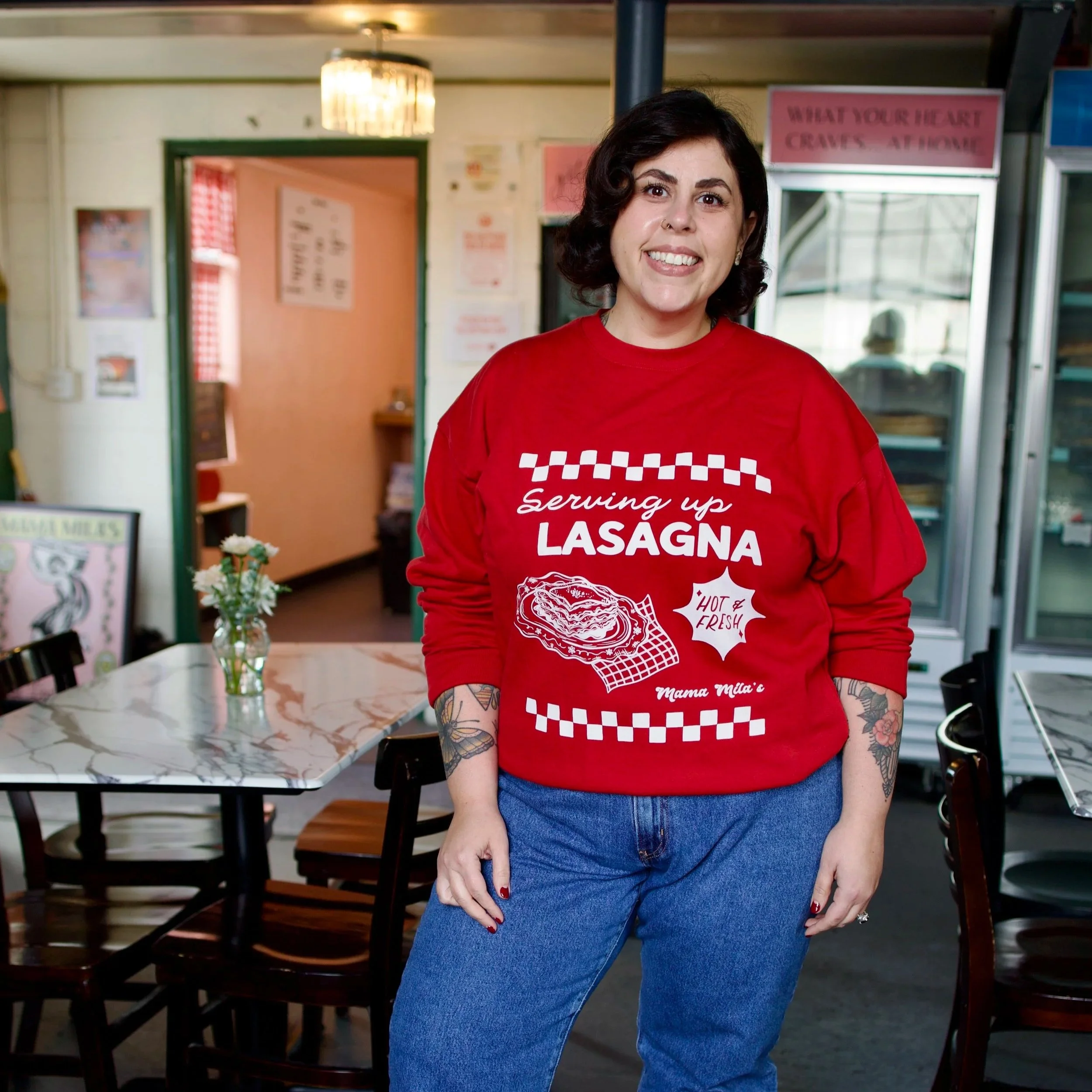 Mila in a red Mama Mila’s sweatshirt featuring a vintage Italian diner-inspired white graphic with “Serving Up Lasagna” lettering, an illustrated plate of lasagna, “Hot & Fresh!” text, and retro check