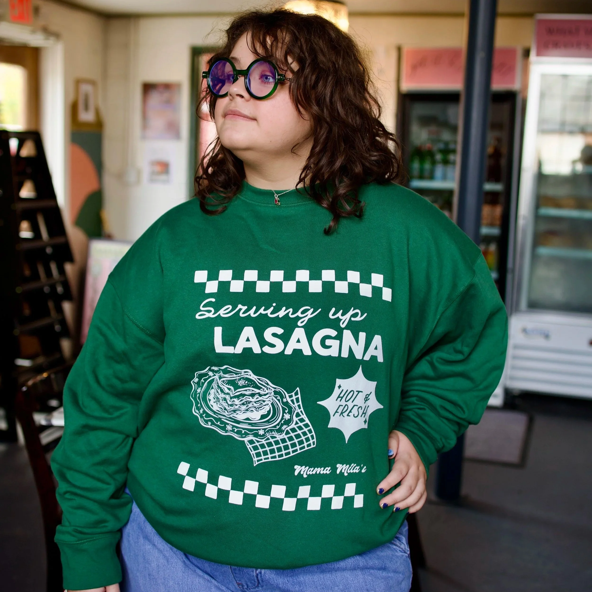 Gia in a green Mama Mila’s sweatshirt featuring a vintage Italian diner-inspired white graphic with “Serving Up Lasagna” lettering, illustrated plate of lasagna, “Hot & Fresh!” text, and retro check