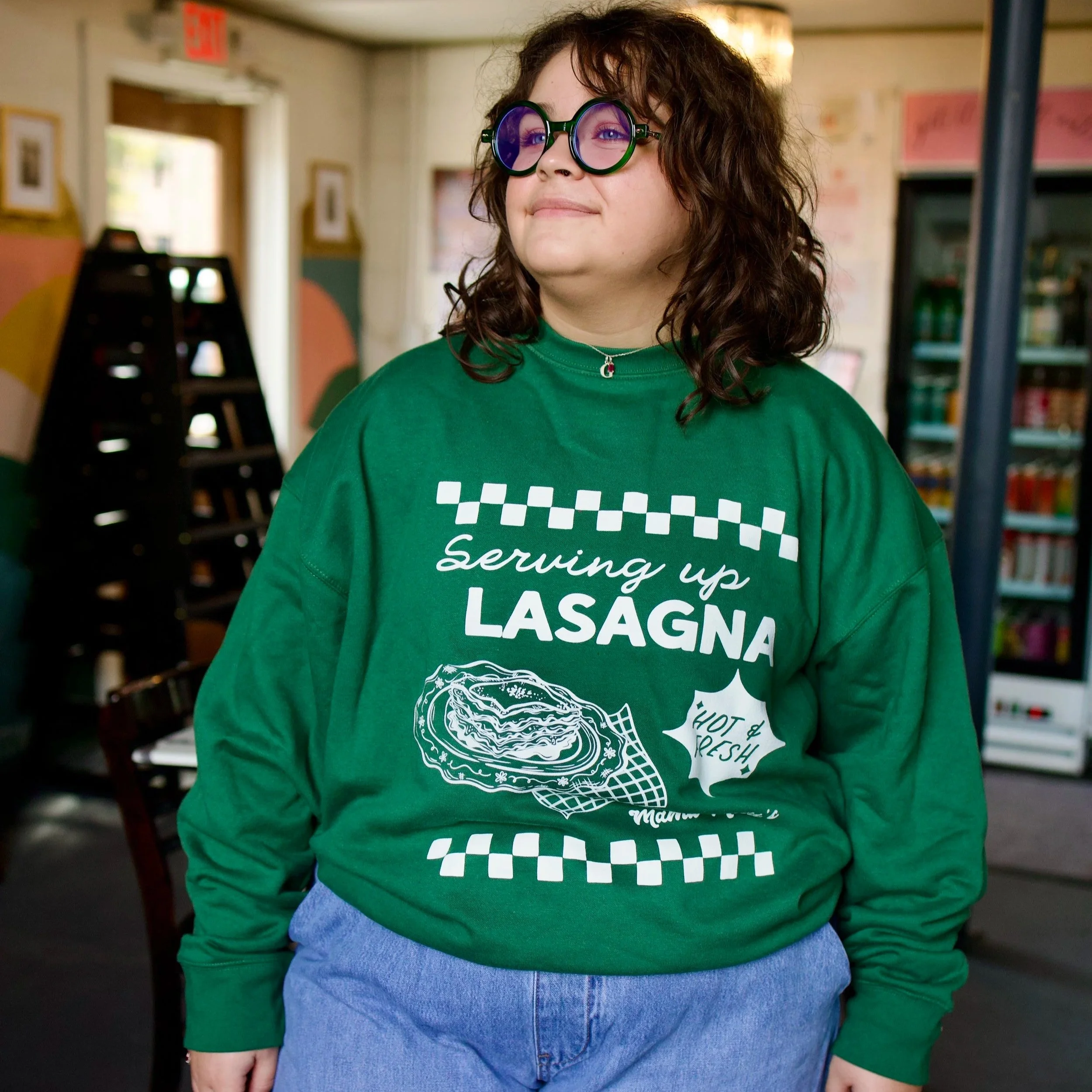 Gia in a green Mama Mila’s sweatshirt featuring a vintage Italian diner-inspired white graphic with “Serving Up Lasagna” lettering, illustrated plate of lasagna, “Hot & Fresh!” text, and retro check