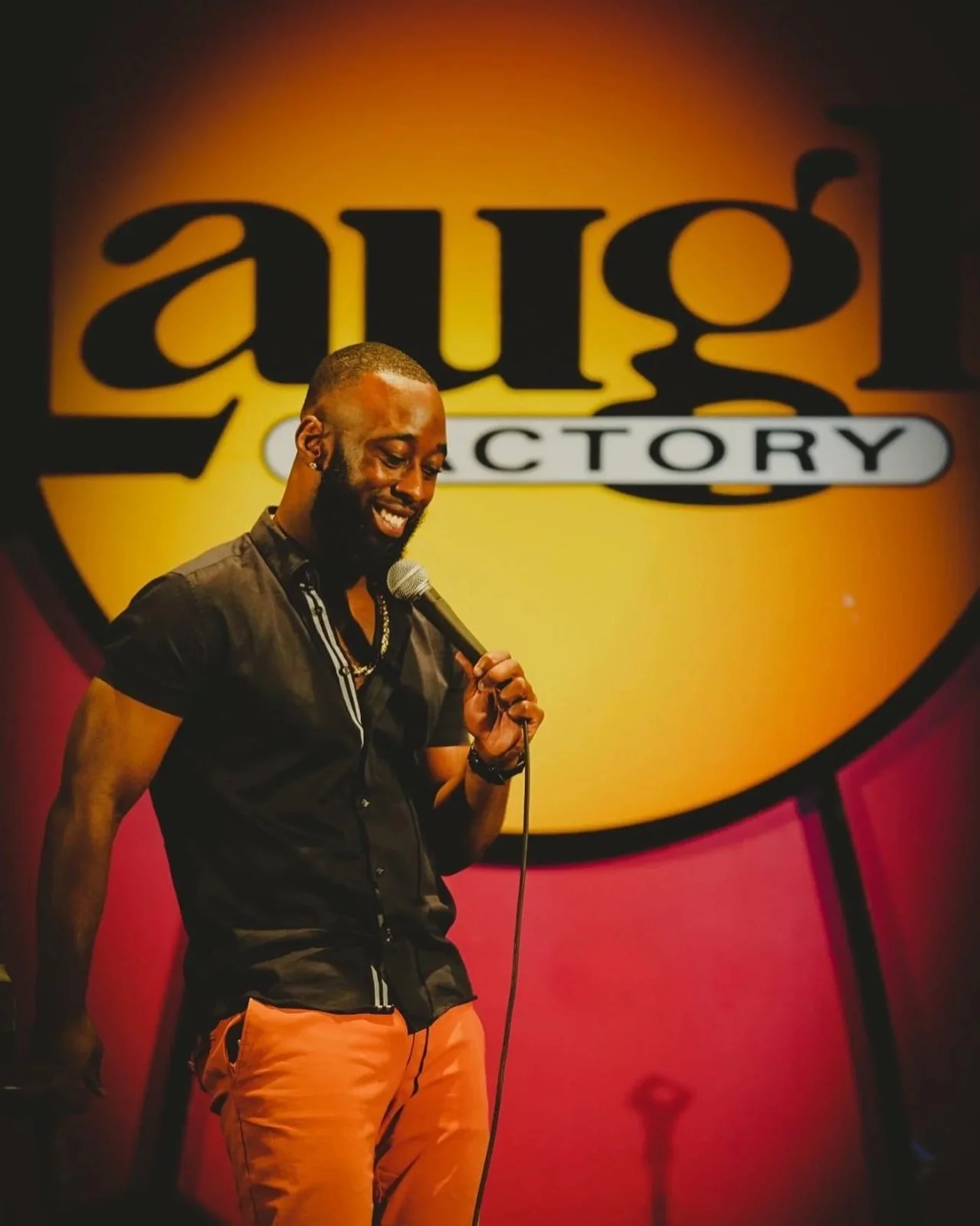St. James Jackson smiling on the Laugh Factory stage in orange trousers.