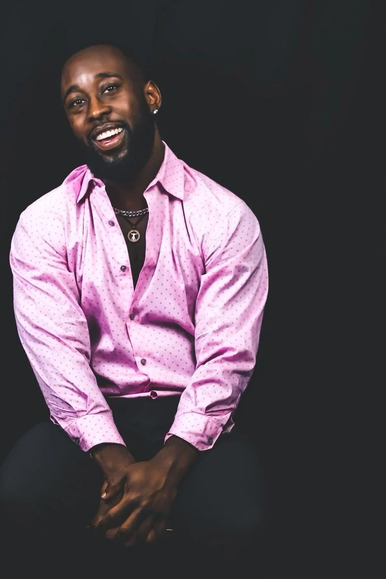 St. James Jackson seated and smiling in a pink shirt against a dark background.