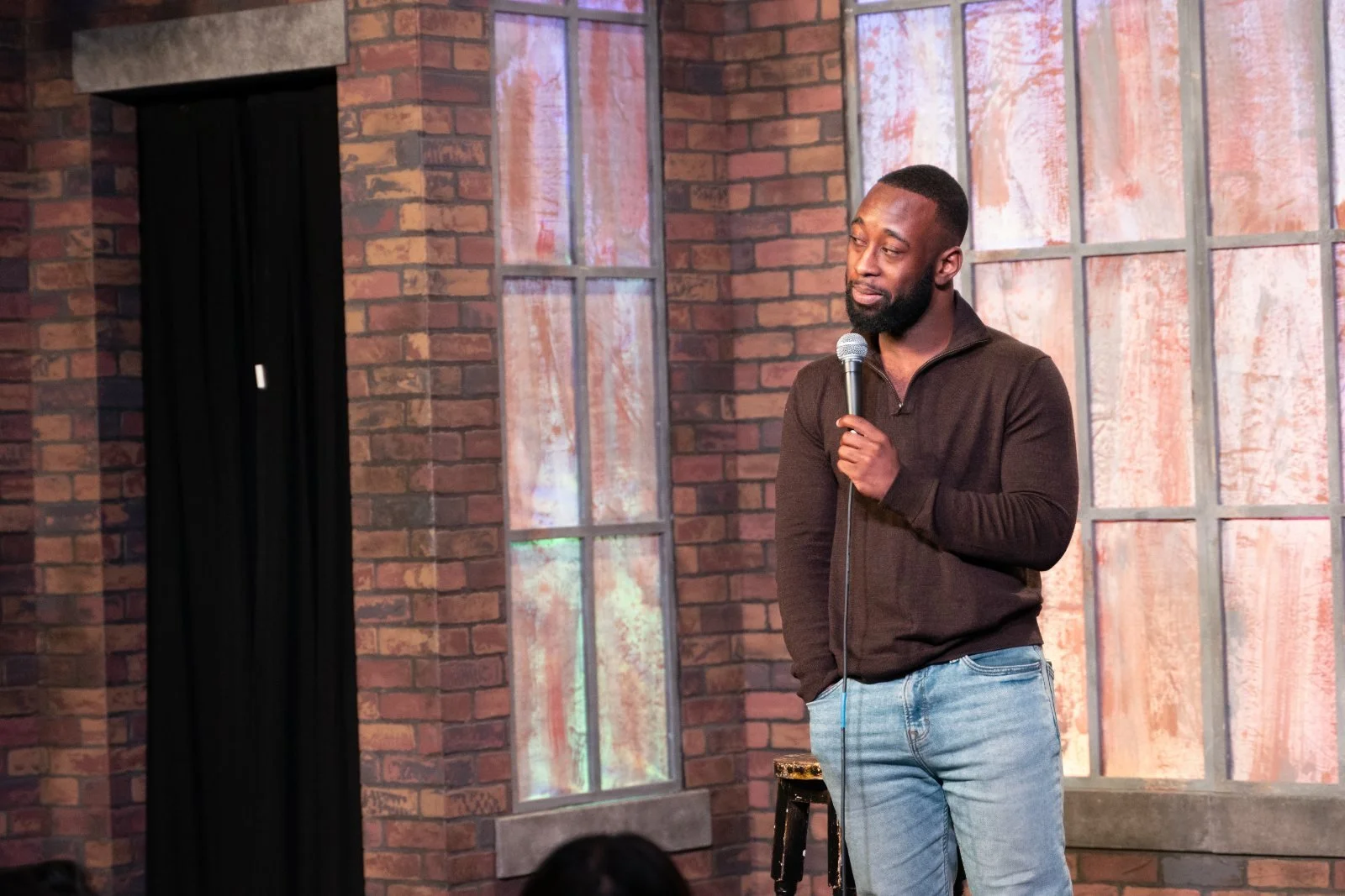 St. James Jackson holding a microphone with one hand in his pocket on a stage with a brick backdrop.