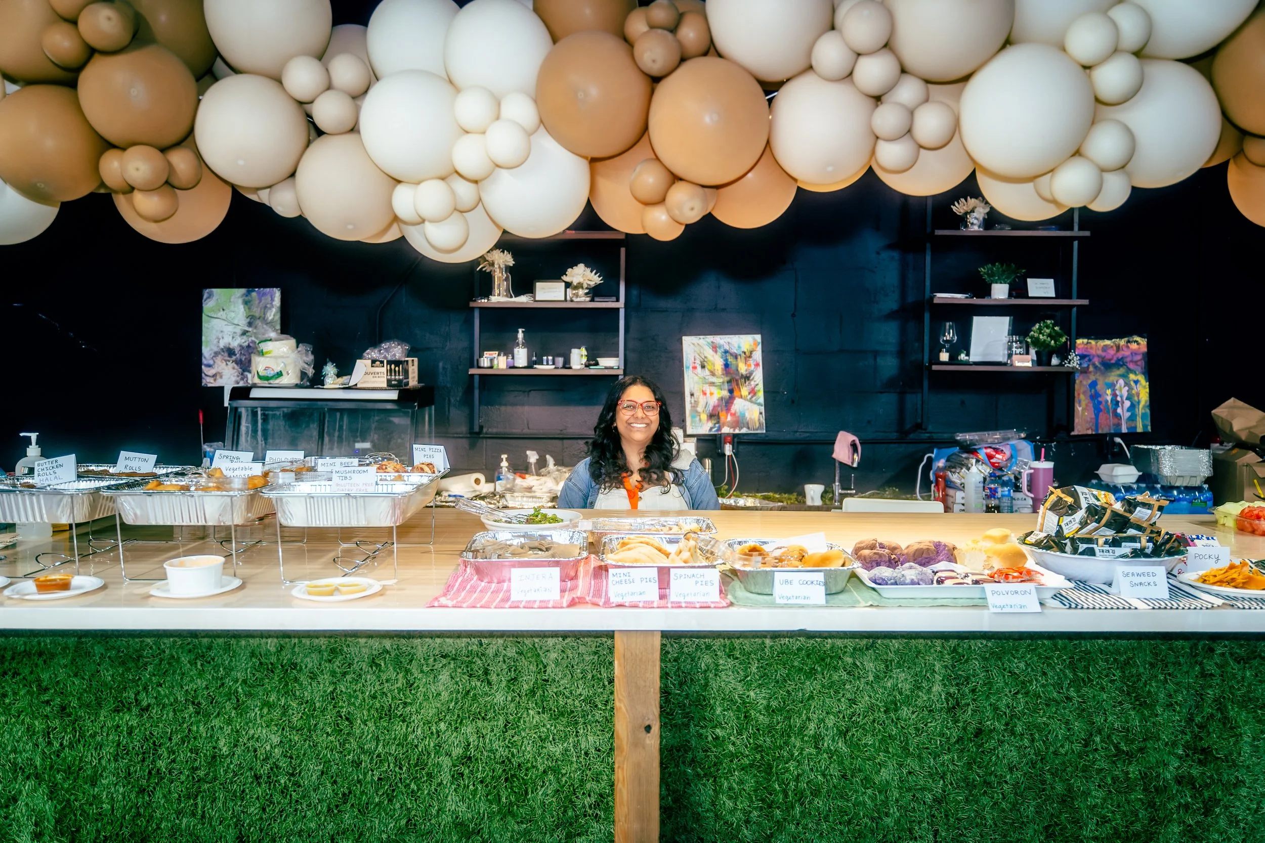 a picture of a person behind all the foods. she is seen smiling straight to the camera wearing a red glass and denim shirt. they are a lot of food in front of her. 