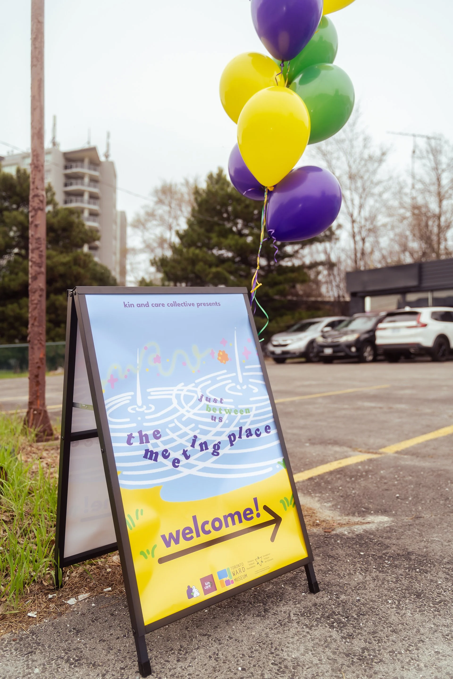 entrance banner that says "kin and care collective presents, just between us, the meeting place. welcome" with arrow to the right showing the way to the event. There are purple and yellow balloon tied on top of the banner.