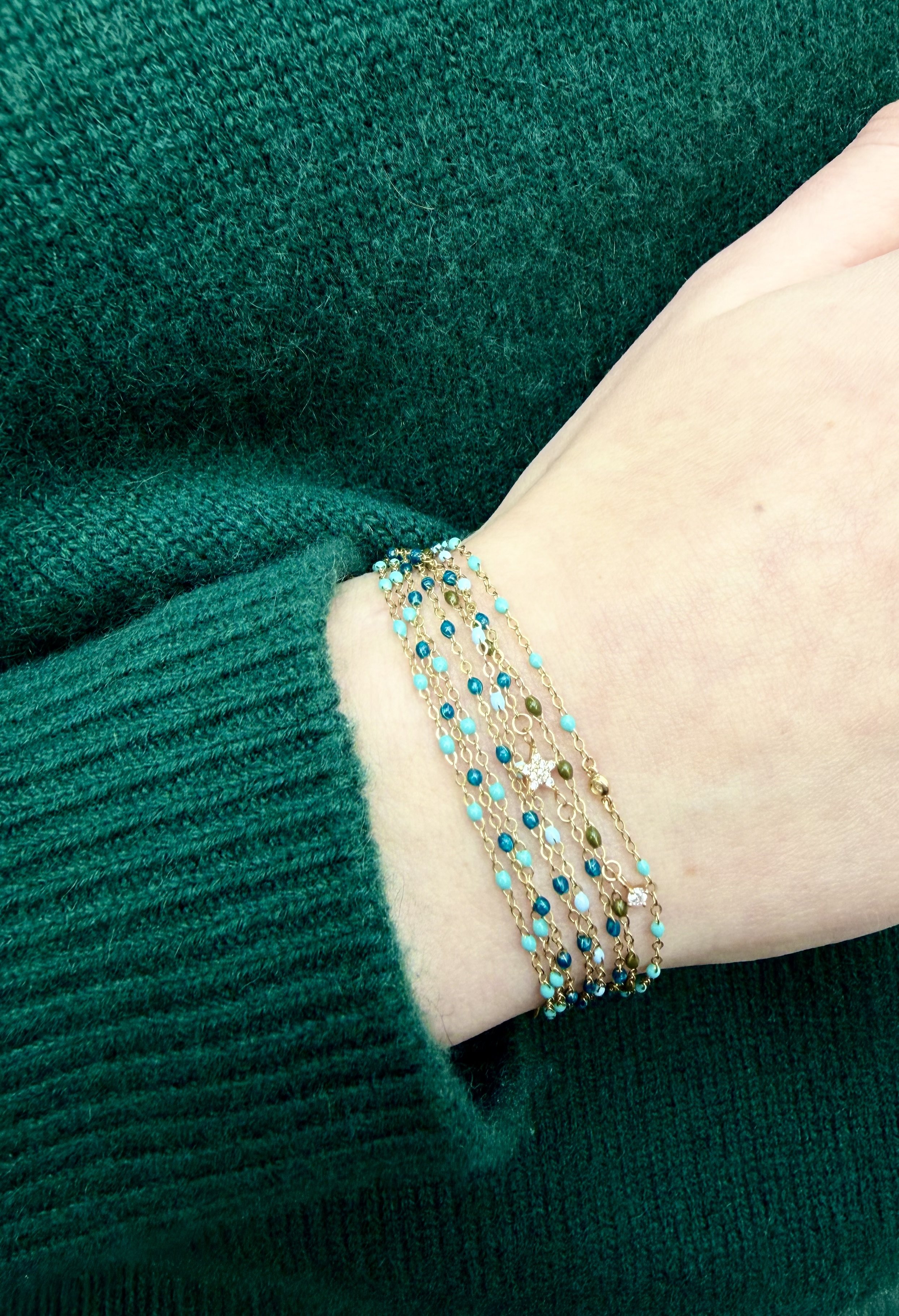 Solid 14k gold enamel chain stack in turquoise tones on wrist, worn over a deep green knit sweater — permanent jewelry by The Sparkle Bar YEG.