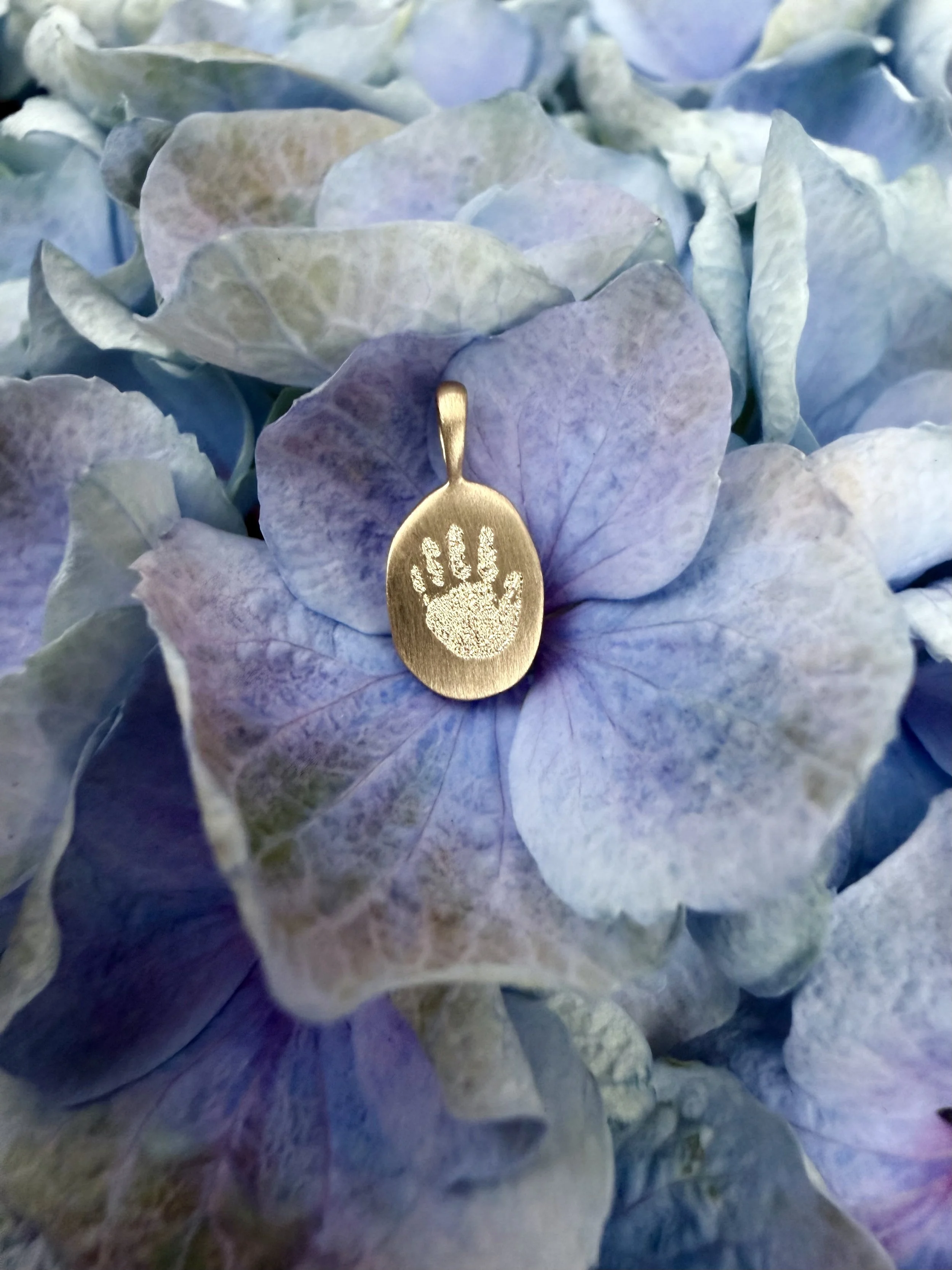 A gold pendant with a handprint engraved on it, resting on blue-hued hydrangea flowers.