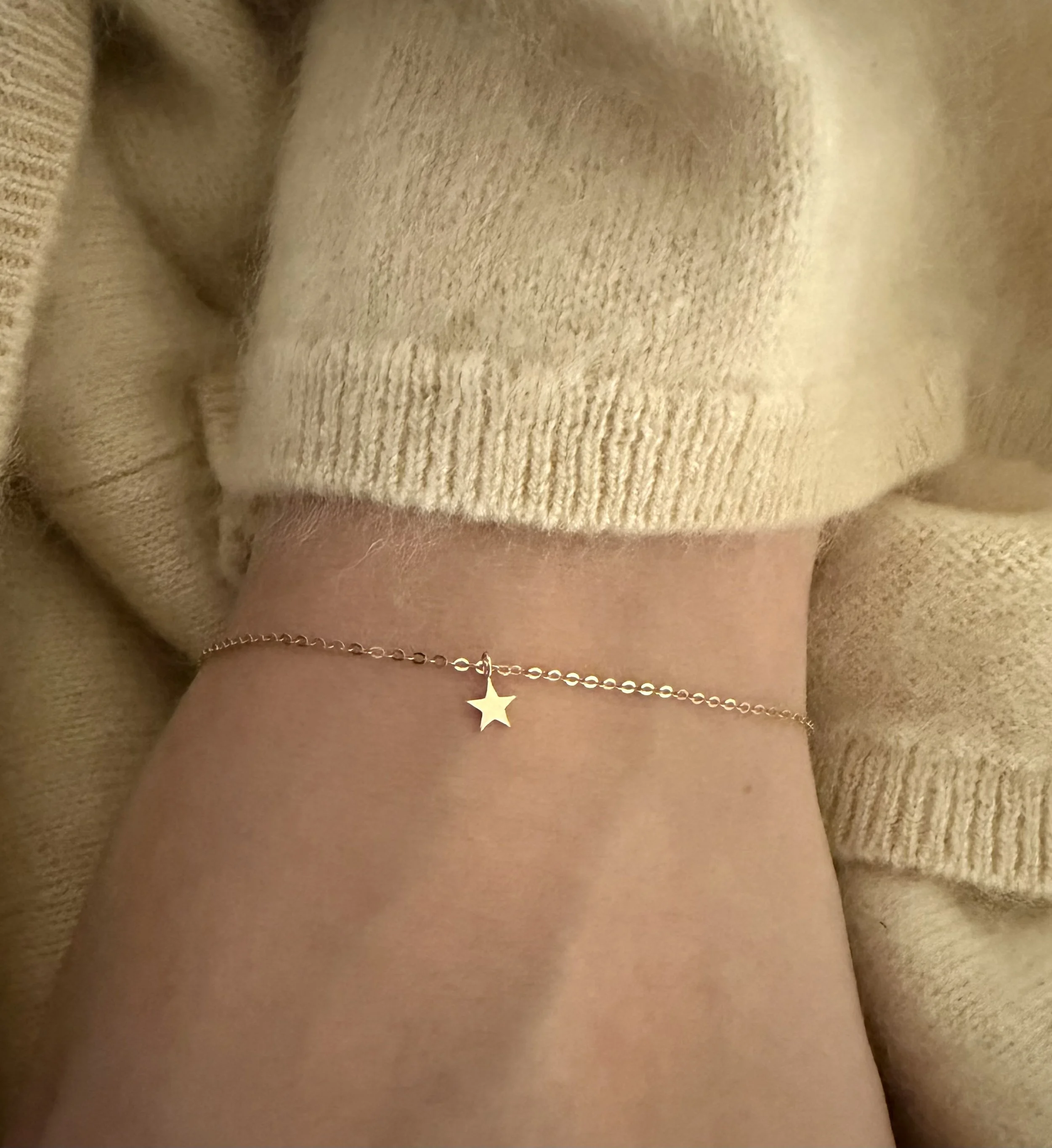 Solid 14k gold star charm bracelet worn on the wrist, styled with a soft beige knit sweater — a delicate permanent jewelry piece from The Sparkle Bar.