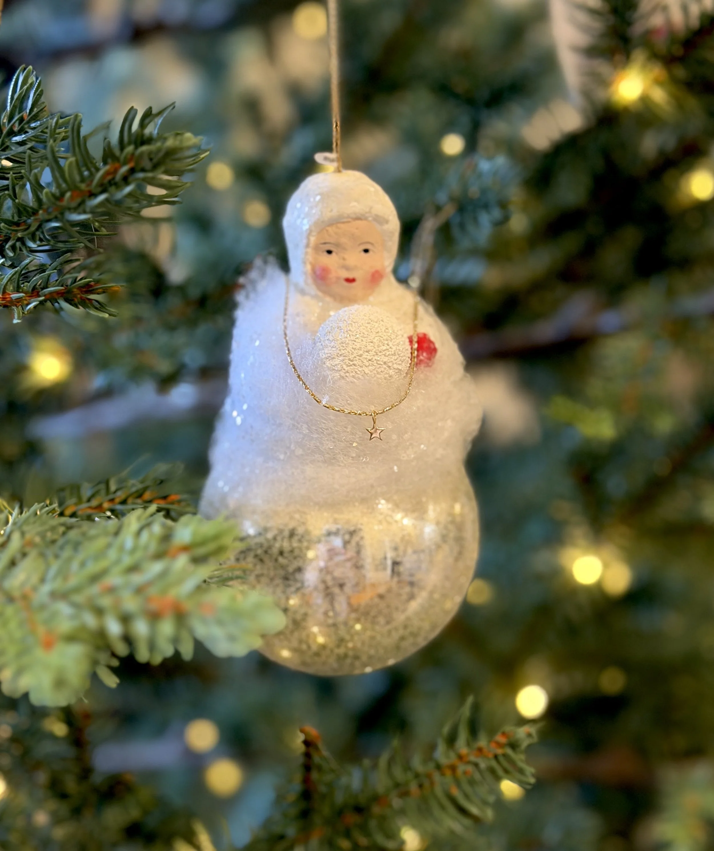 Vintage snow-baby ornament on a Christmas tree wearing a delicate solid 14k gold star charm necklace from The Sparkle Bar, softly lit with warm holiday lights.