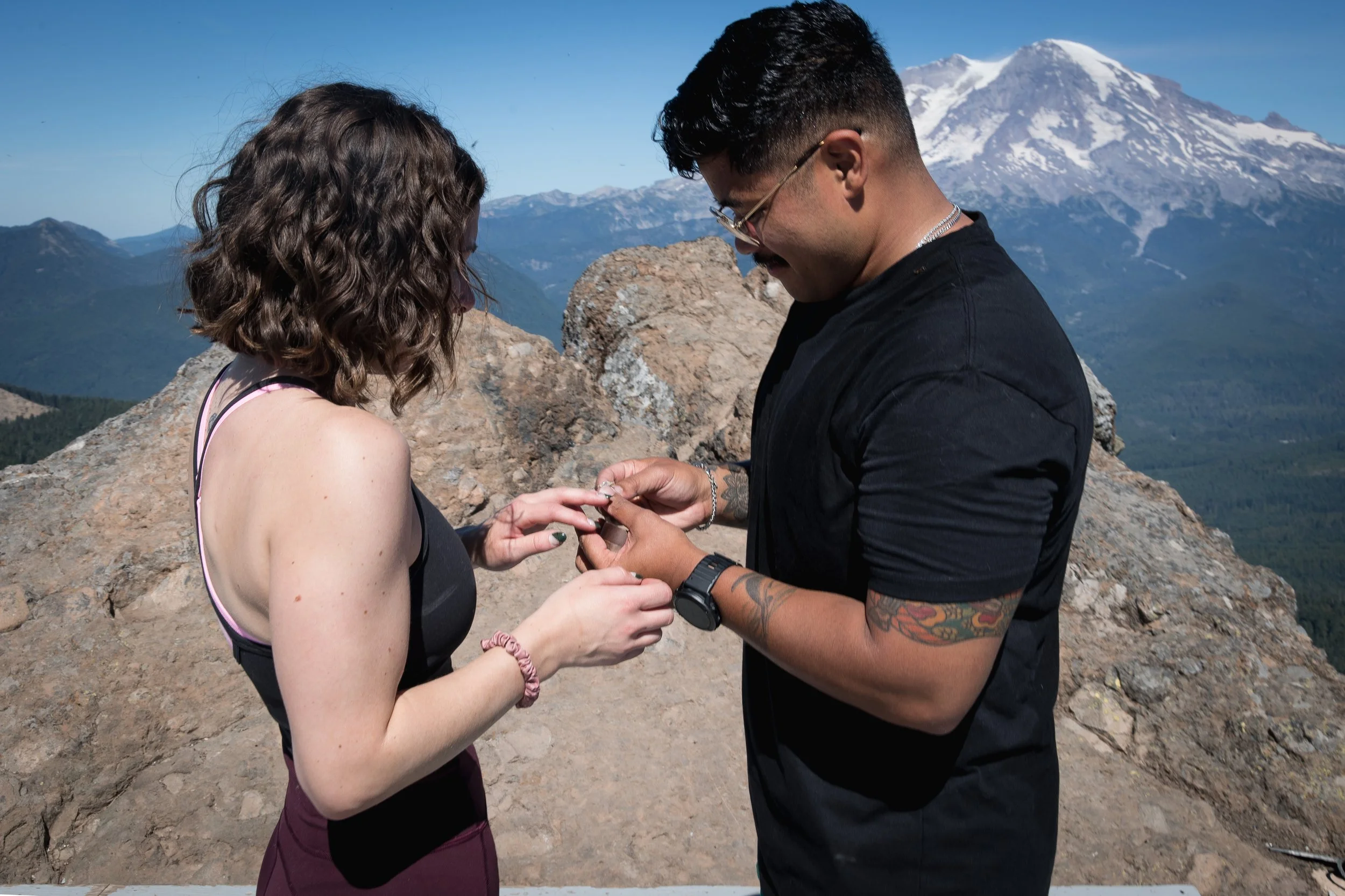 A man is proposing marriage to a woman on top of a mountain with a snow-capped Mt. Rainier in the background.