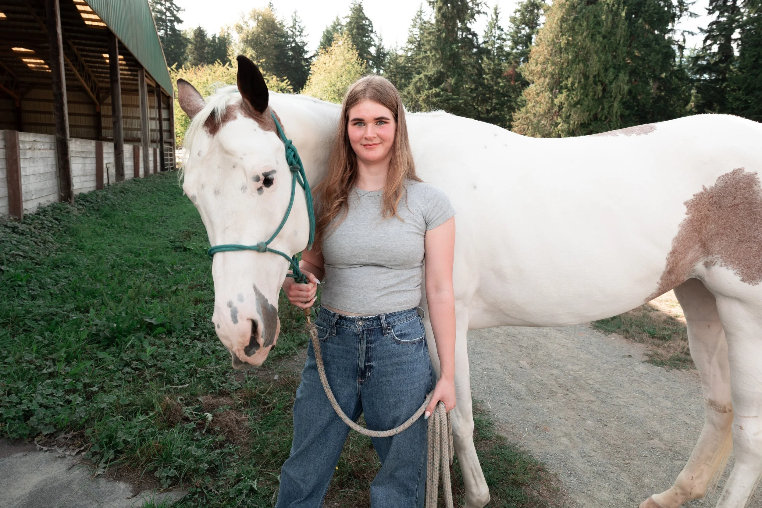 A young woman with long brown hair, wearing a gray t-shirt and jeans, stands outdoors holding a lead rope connected to a white horse with brown spots and a teal halter. The horse is leaning its head toward her, and there are trees and a building structure in the background.
