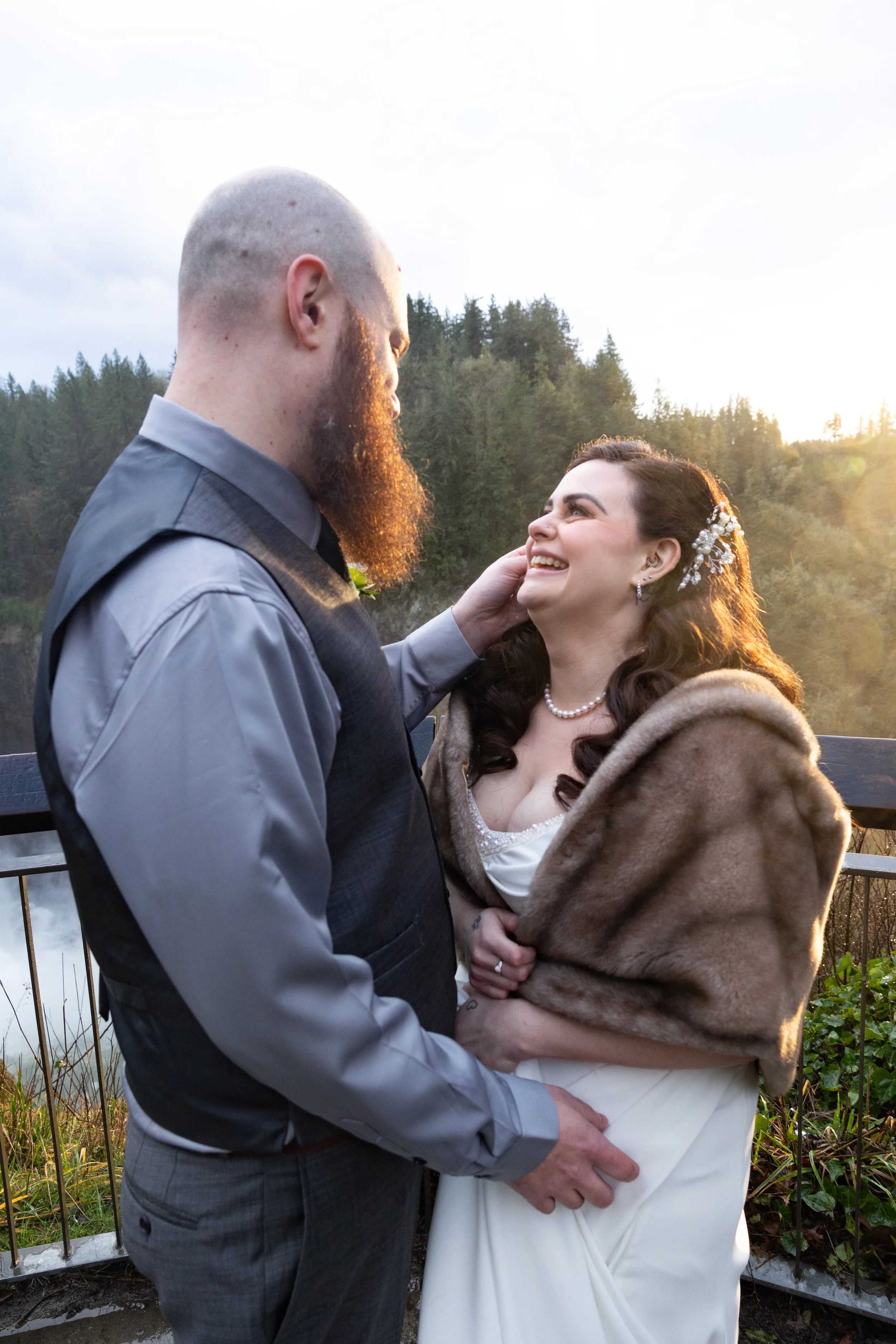 A bride and groom standing outdoors, facing each other and smiling. The groom has a beard and is dressed in a gray shirt and vest, while the bride wears a fur stole over her wedding dress and has a pearl necklace.