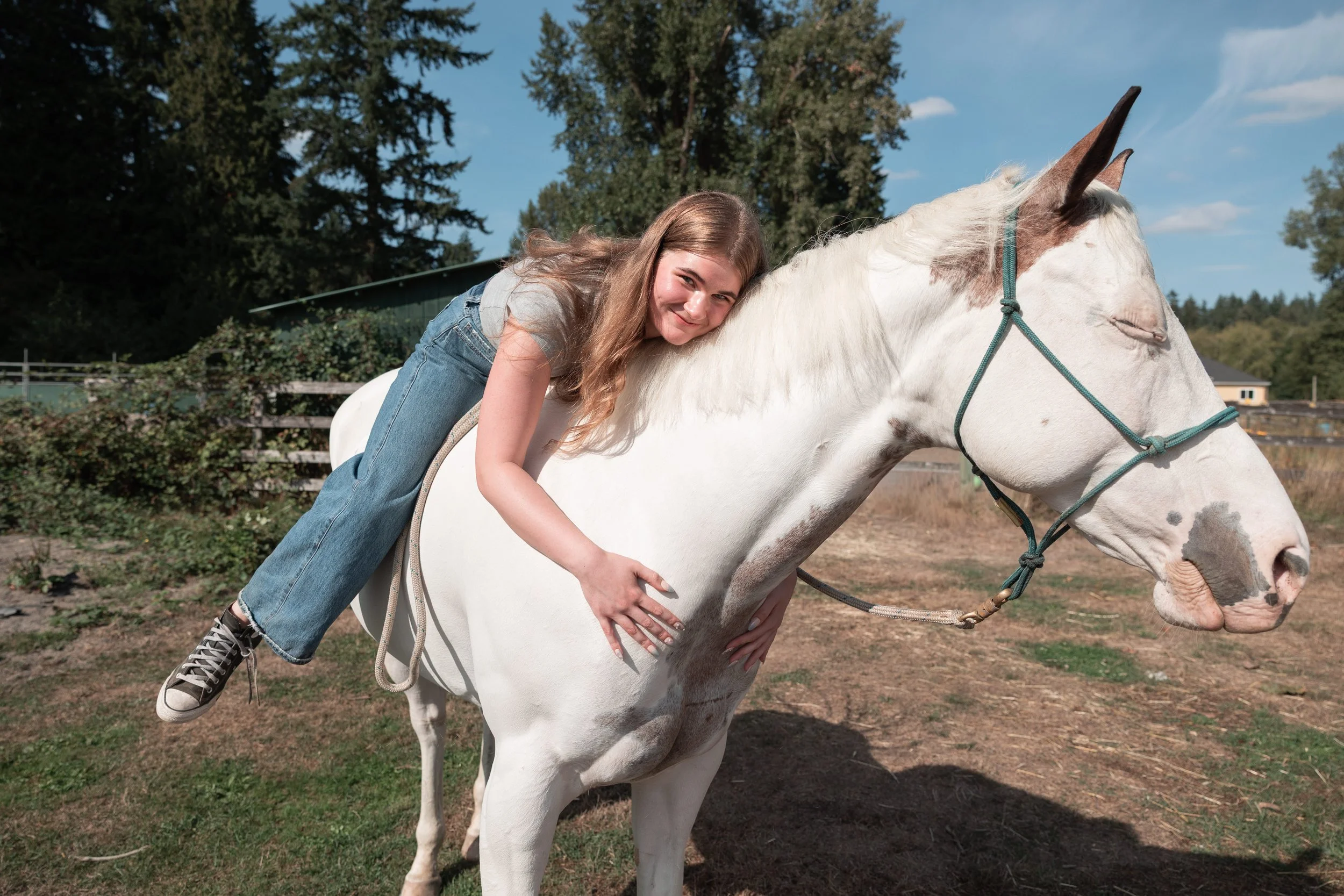 A young woman with long brown hair smiling while lying on the neck of a white horse with brown markings on its face and ears, outdoors on a sunny day with trees and a fence in the background.