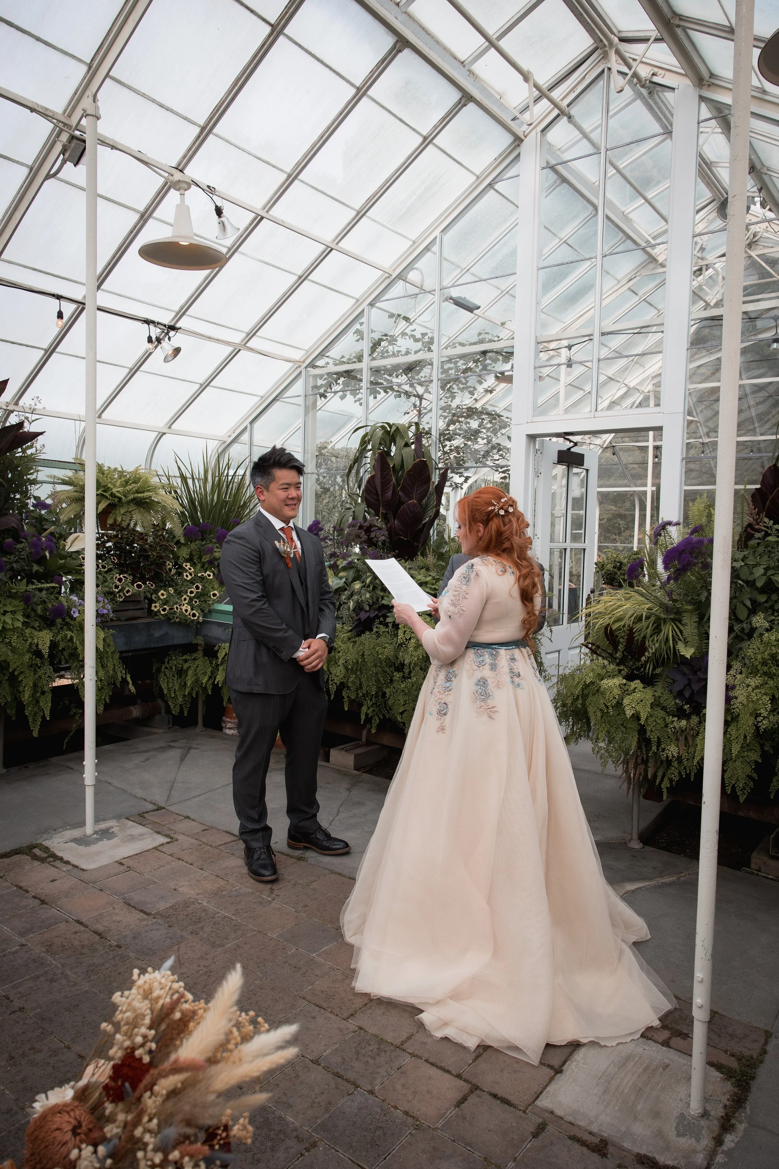 A bride and groom exchange vows in a greenhouse filled with lush plants and flowers during a wedding ceremony at Volunteer Park Conservatory.