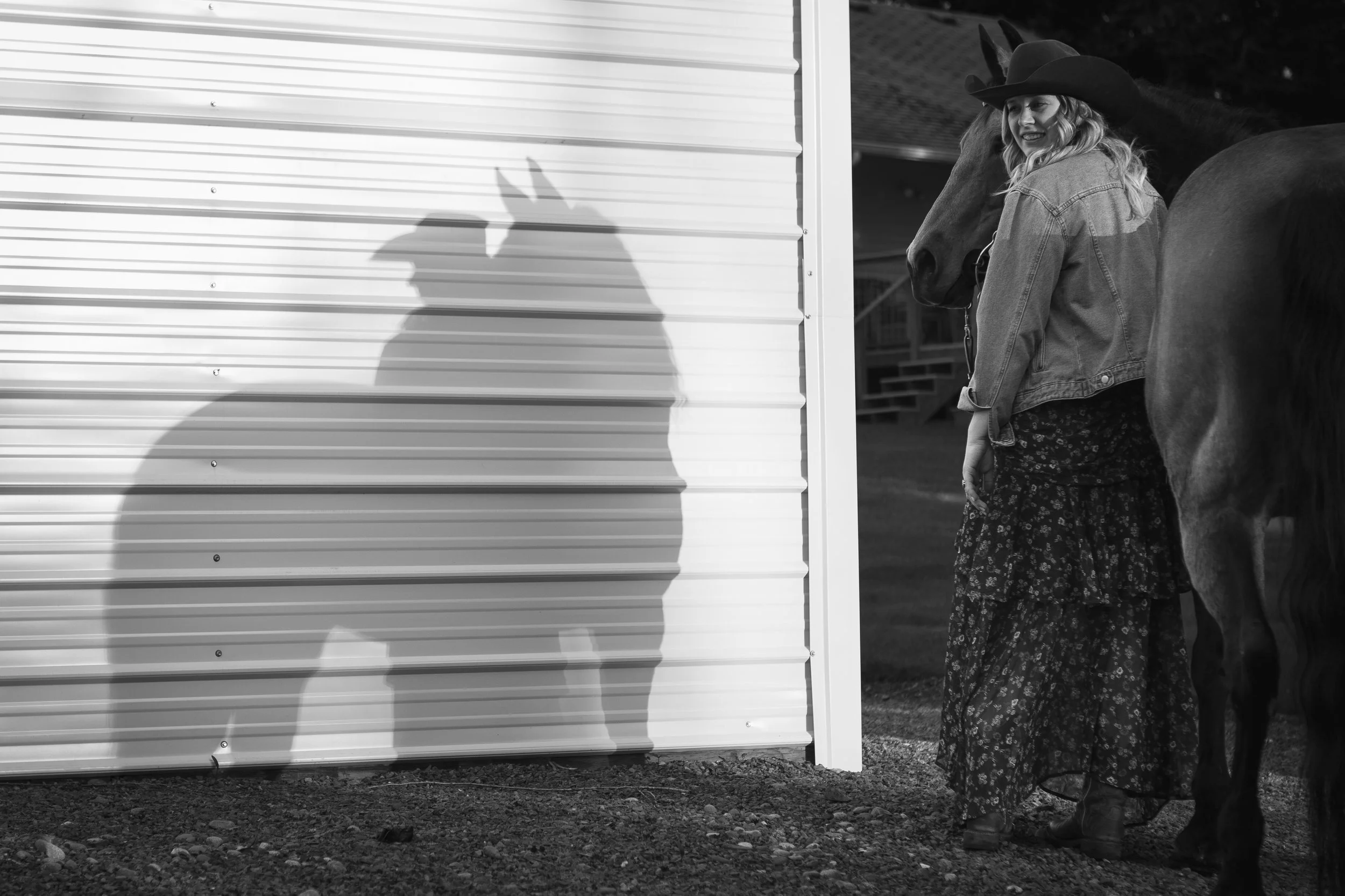 A woman dressed in a long skirt, denim jacket, and cowboy hat stands beside a gray horse, with her candid smile visible. Her shadow is cast on a white metal wall beside her, with the horse's face almost touching her.