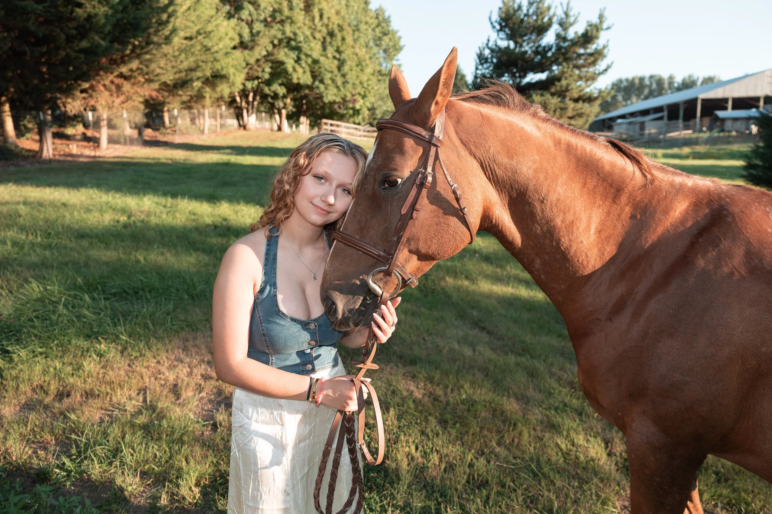 A young woman with curly blonde hair, wearing a denim vest and white skirt, standing next to a brown horse with a bridle, on a grassy field with trees and farm buildings in the background.