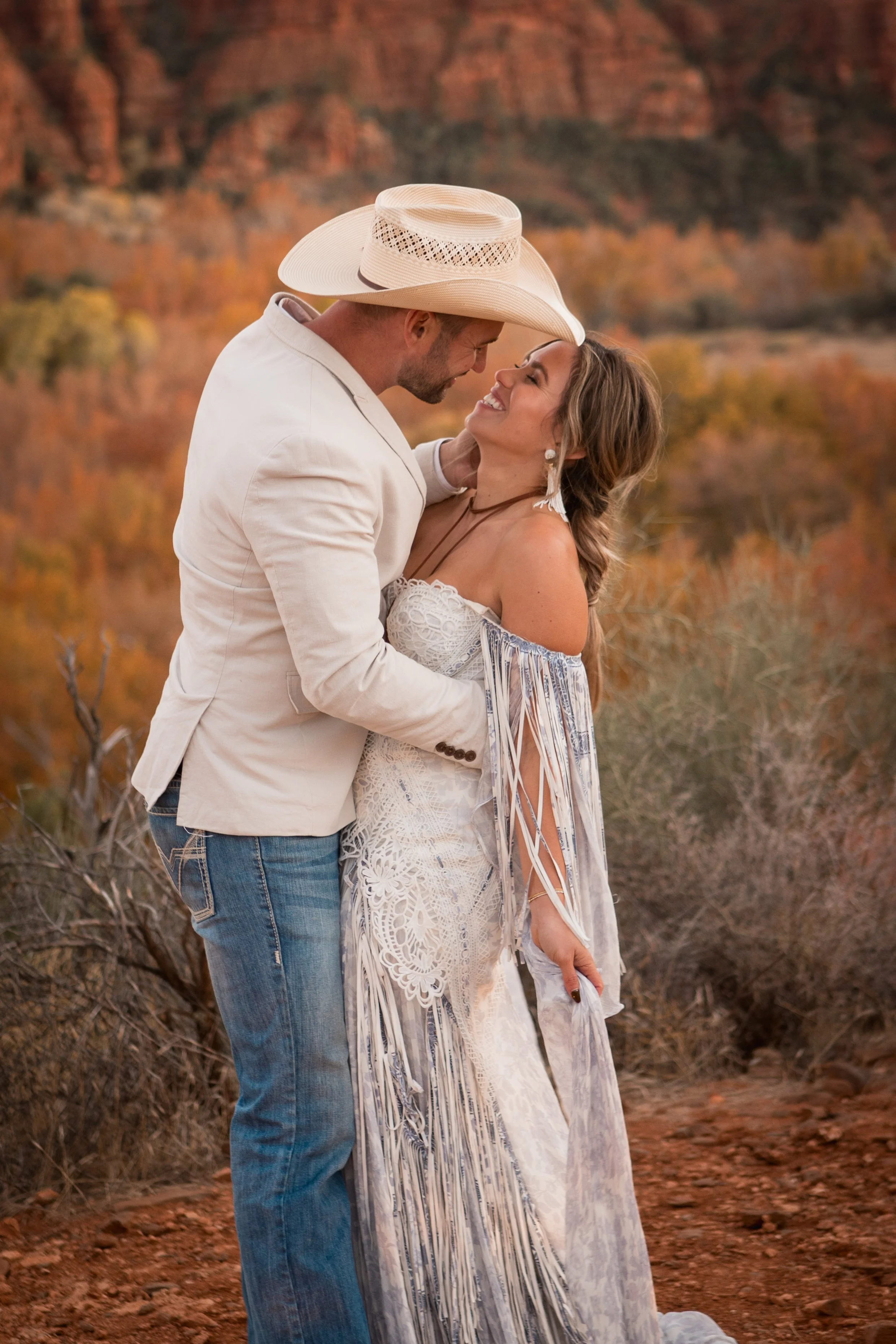 A couple smiling and looking at each other in a romantic embrace outdoors with autumn fall foliage in the background.