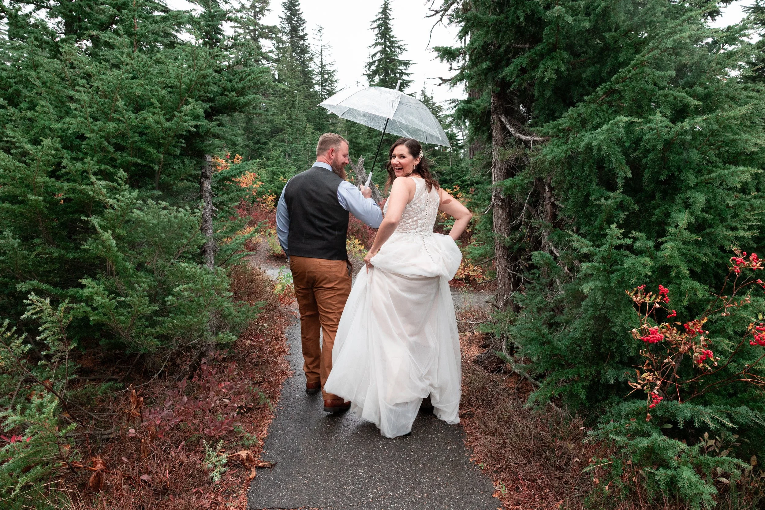 A bride and groom walking on a forest trail, sharing a laugh under a transparent umbrella, surrounded by tall green trees and colorful plants.