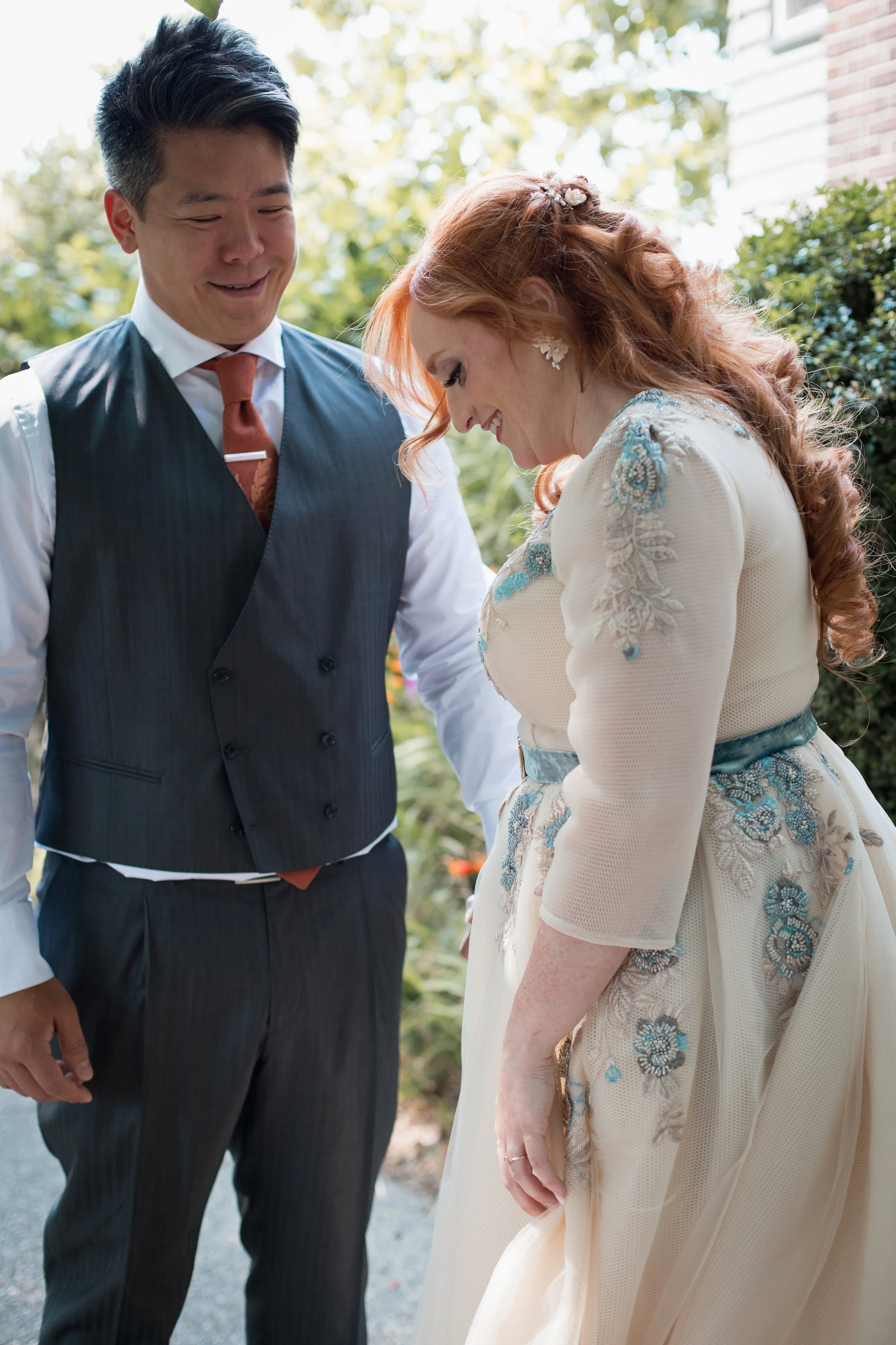 A couple sharing a joyful moment outdoors, dressed in wedding attire, with the woman in a cream-colored gown with blue floral embroidery and the man in a dark vest and red tie, smiling and looking down.