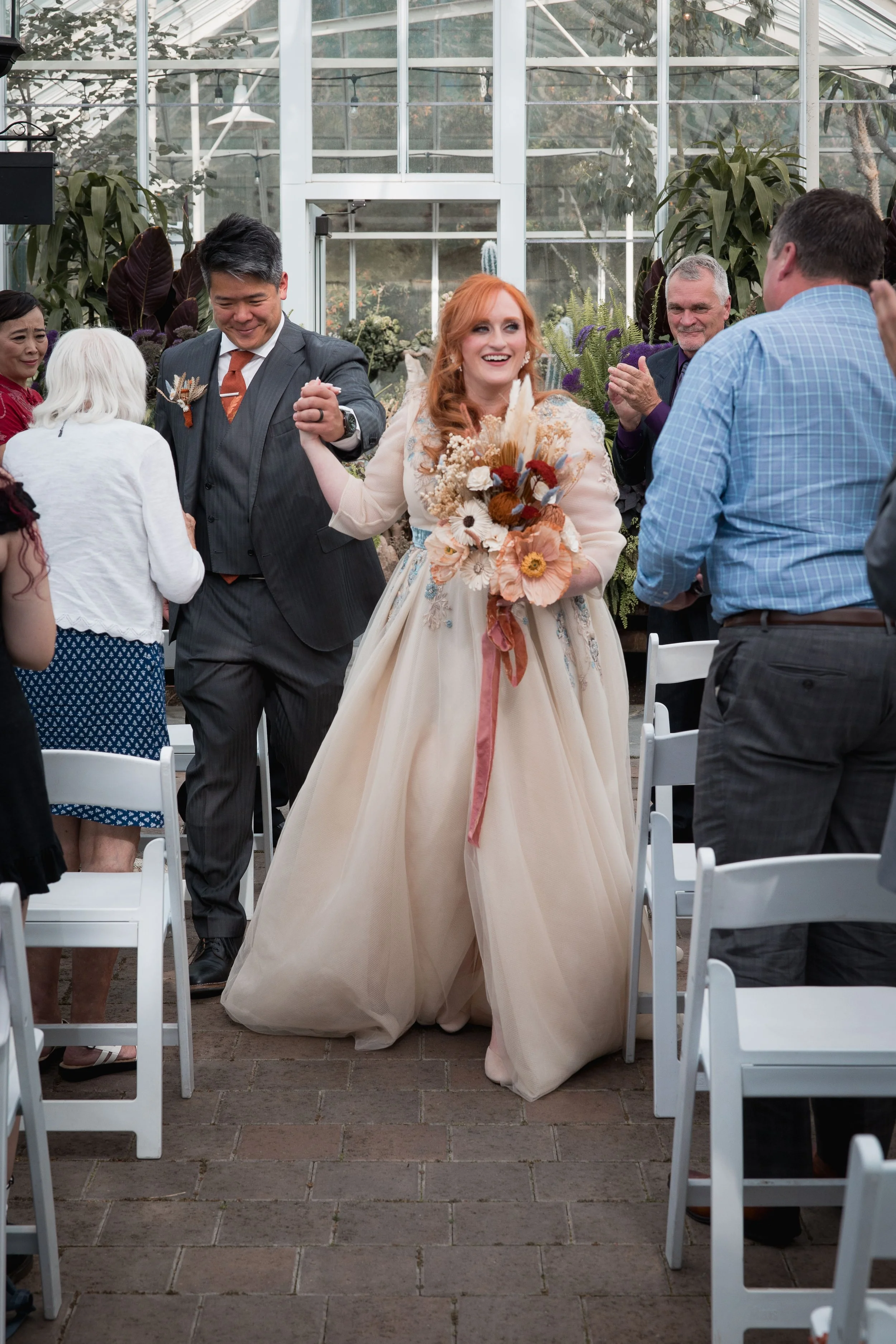 A bride with red hair holding a bouquet of flowers walking down the aisle at a wedding ceremony, surrounded by guests in a greenhouse setting.