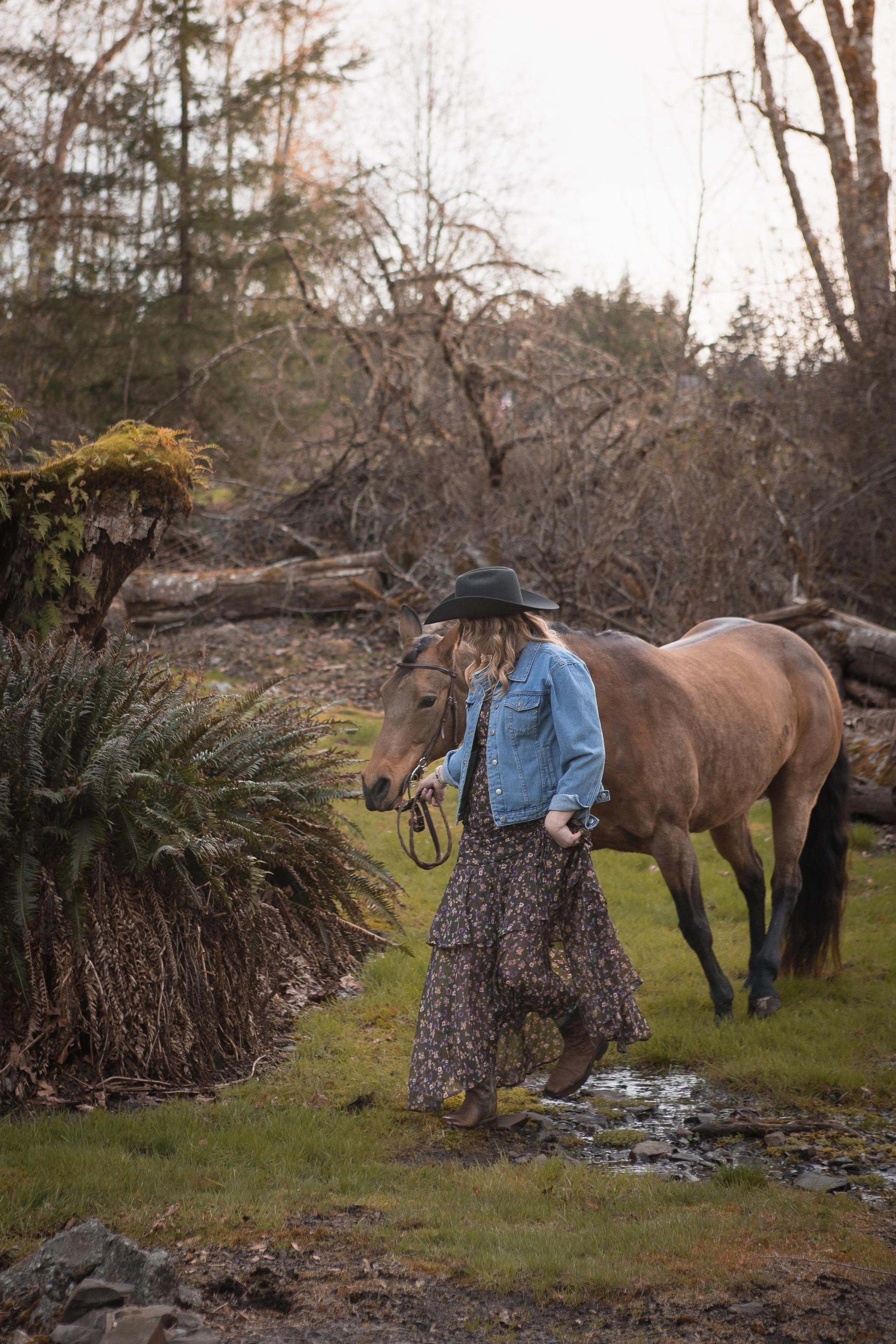 A woman with long blonde hair, wearing a black hat, denim jacket, floral dress, and boots, leading a light brown horse across a small stream in a wooded area during the daytime.